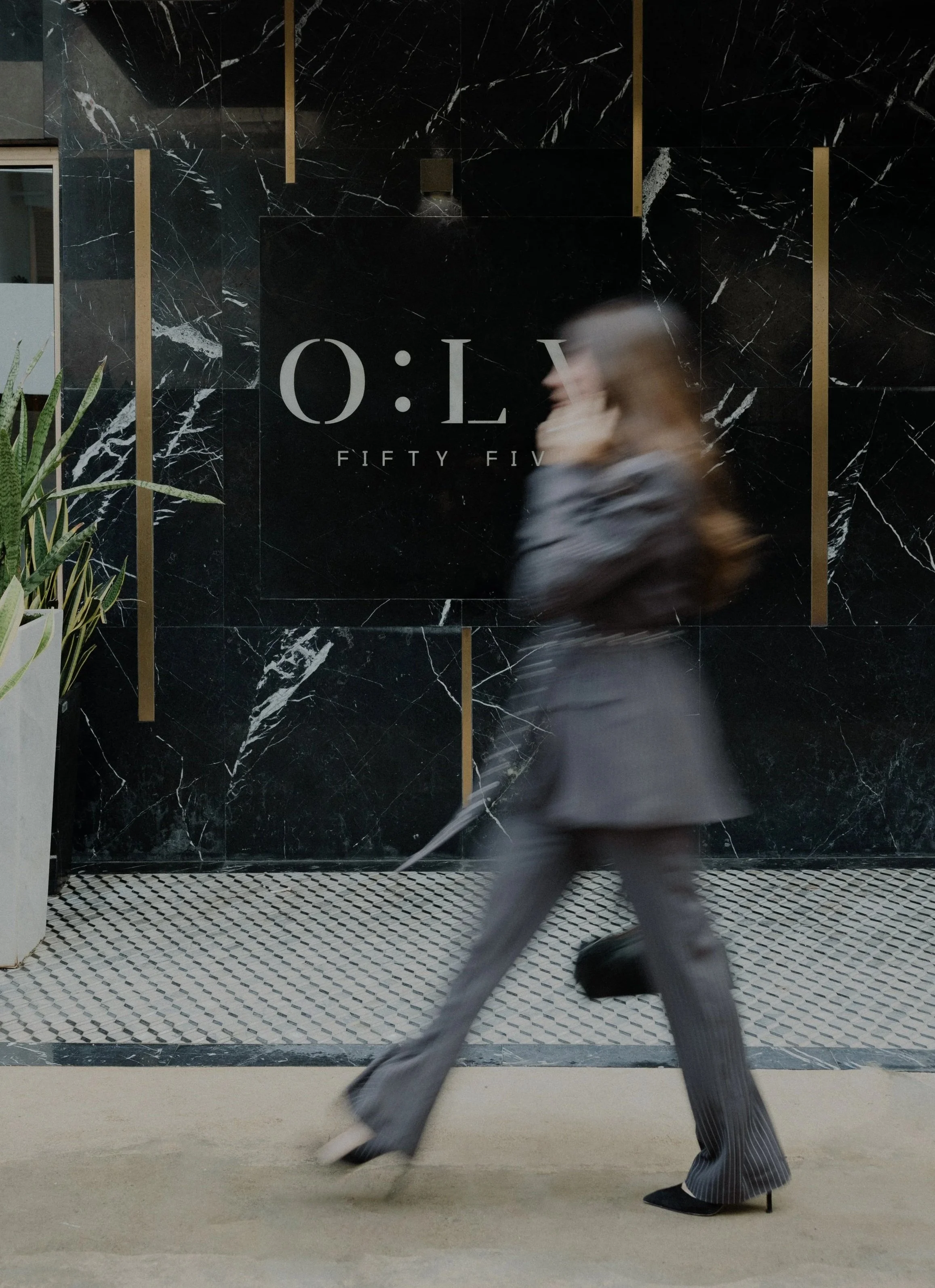 A woman with shoulder-length blonde hair, dressed in a gray top and pinstriped pants, walking quickly in front of a black marble wall with the text 'FIFTY FIVE' and 'O:L' on it.