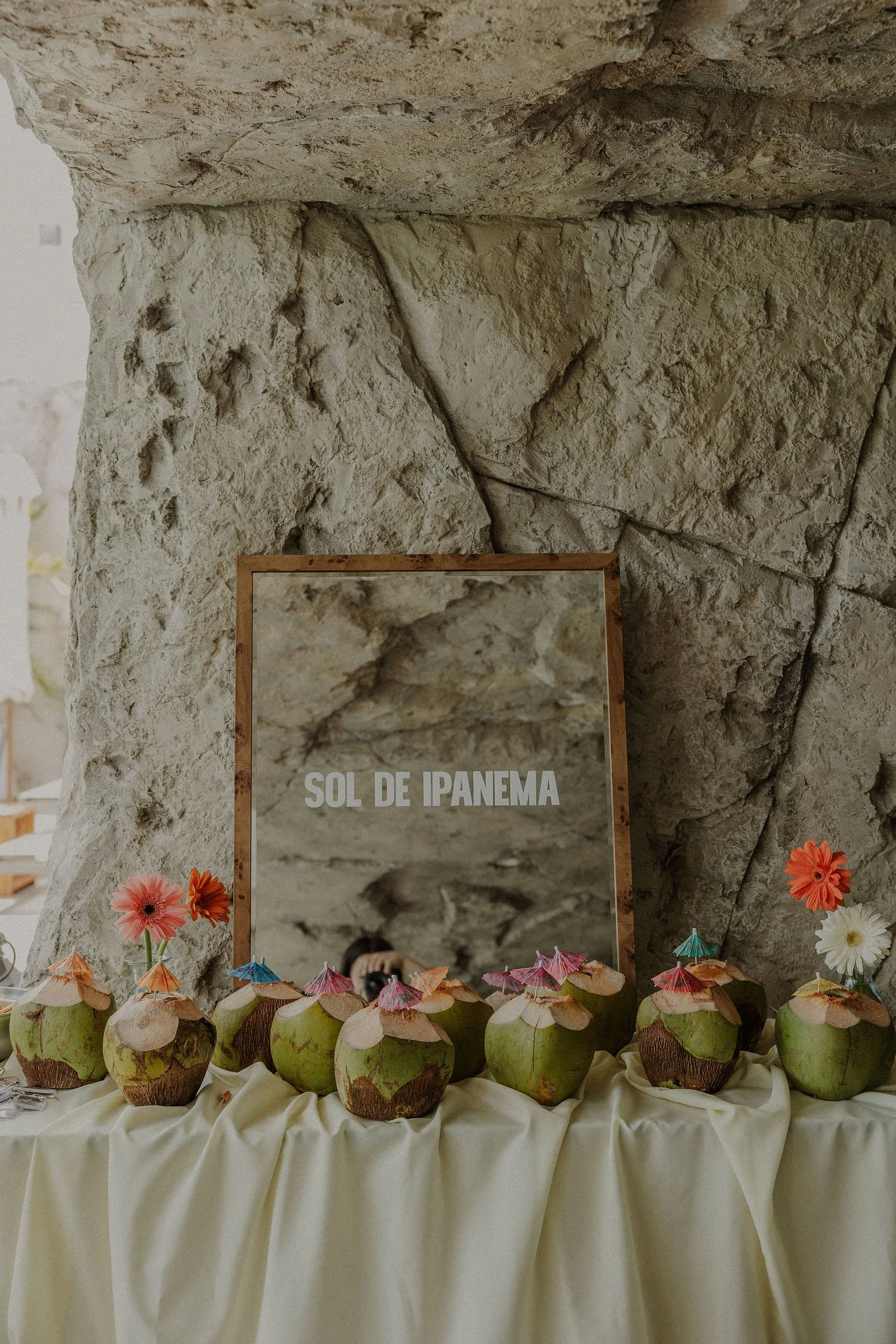 A display of fresh coconuts decorated with miniature paper umbrellas and flowers, arranged on a table covered with a cream-colored cloth, with a rocky wall and a mirror behind them. The mirror has the text "Sol de Ipanema."