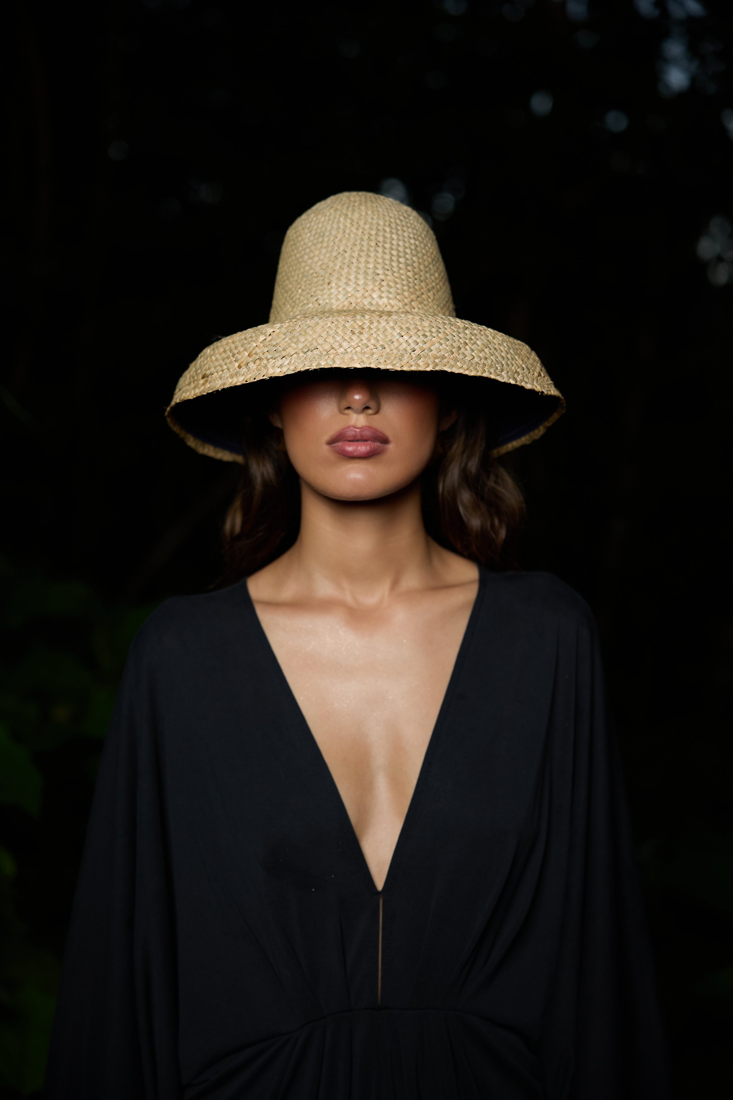 A woman wearing a wide-brimmed straw hat with her face partially obscured, dressed in a black V-neck dress, standing outdoors against a dark background.