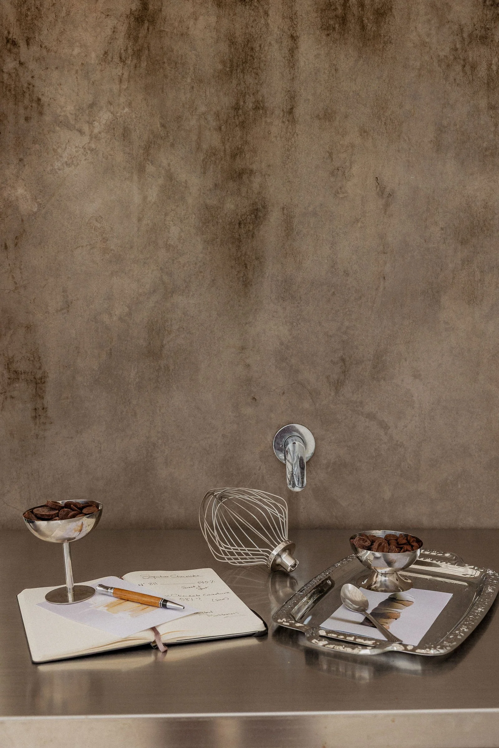 A kitchen counter with an open notebook and pen, two bowls of chocolate candies on stands, a wire whisk, a silver tray with spoons and a dessert with chocolate and spoons. The background has a textured wall with a metallic faucet.
