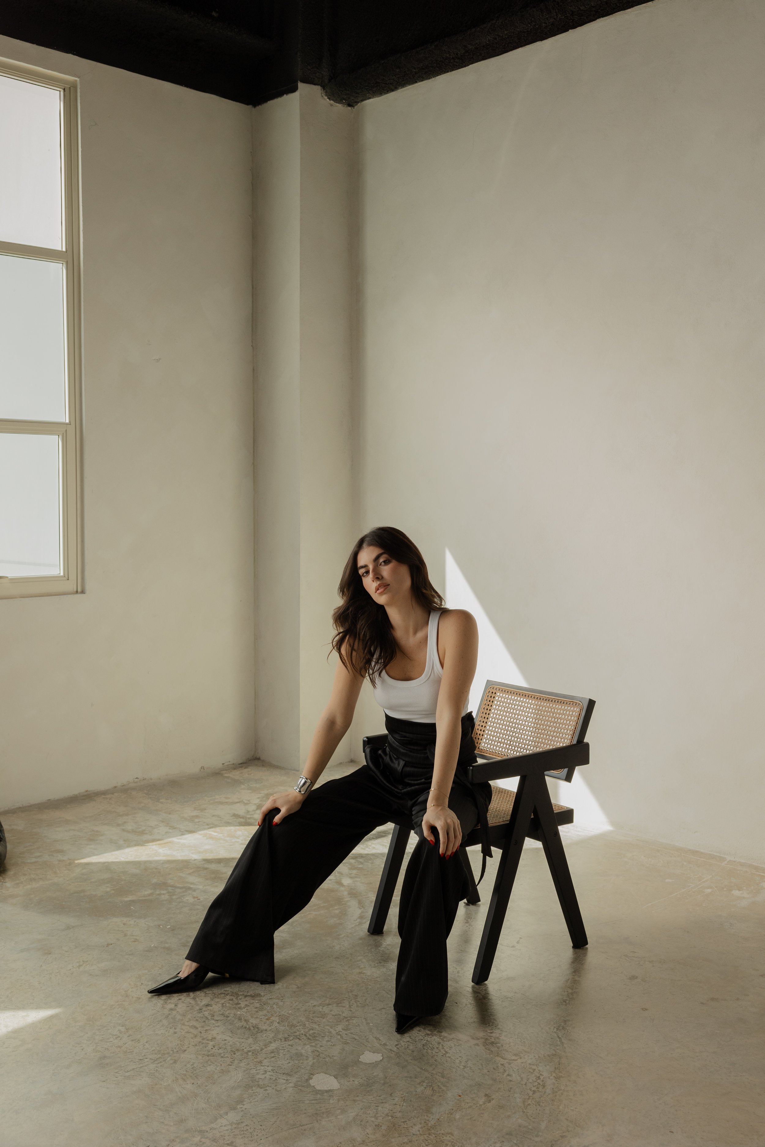 A woman with long wavy hair dressed in a white top and black wide-leg pants is sitting on a chair in a minimalist room with white walls, a concrete floor, a window, and natural light casting shadows.