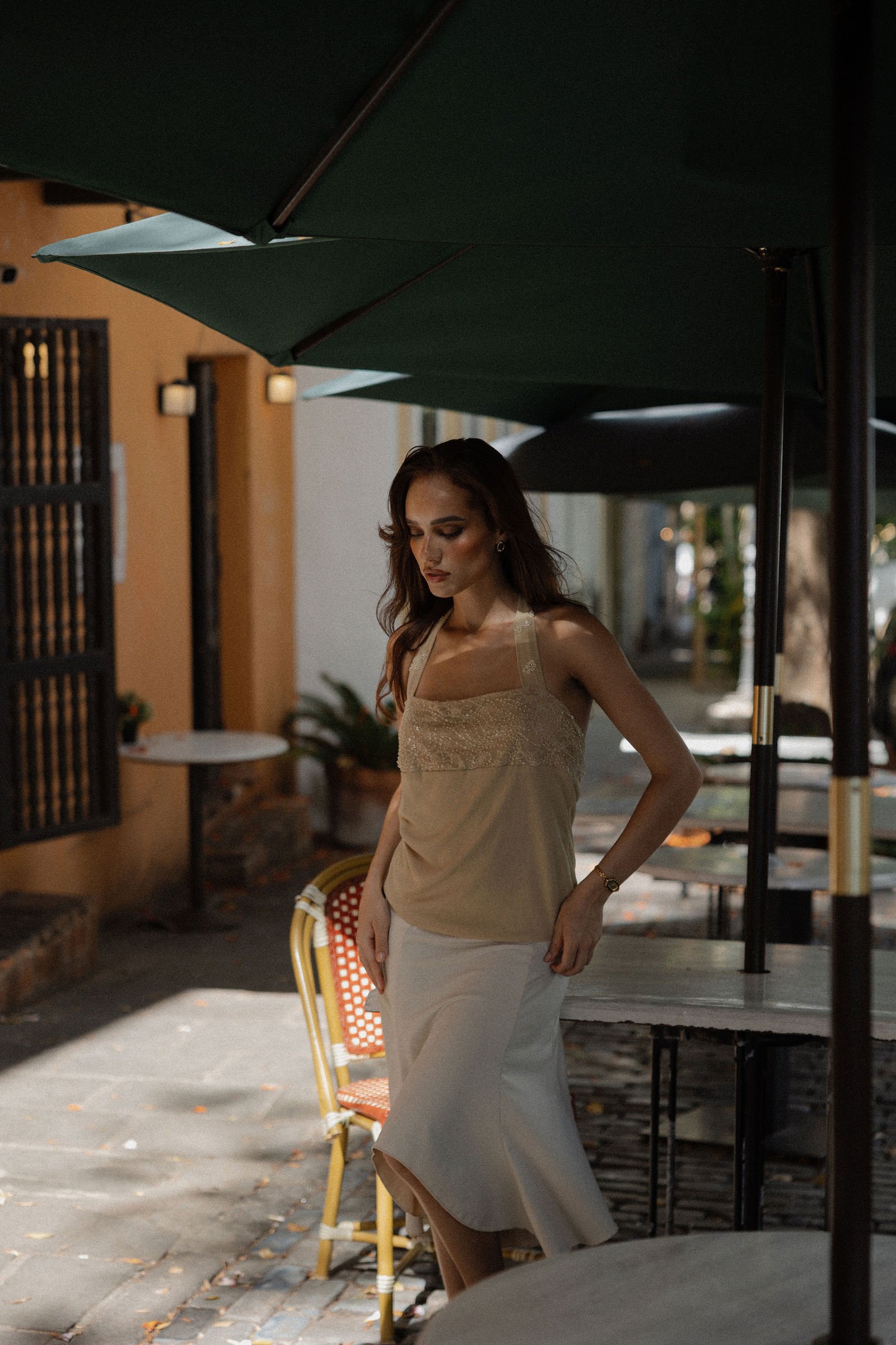 A woman with red hair and earrings wearing a beige top and a white skirt, standing outdoors under a dark green umbrella, with closed eyes and a neutral expression.