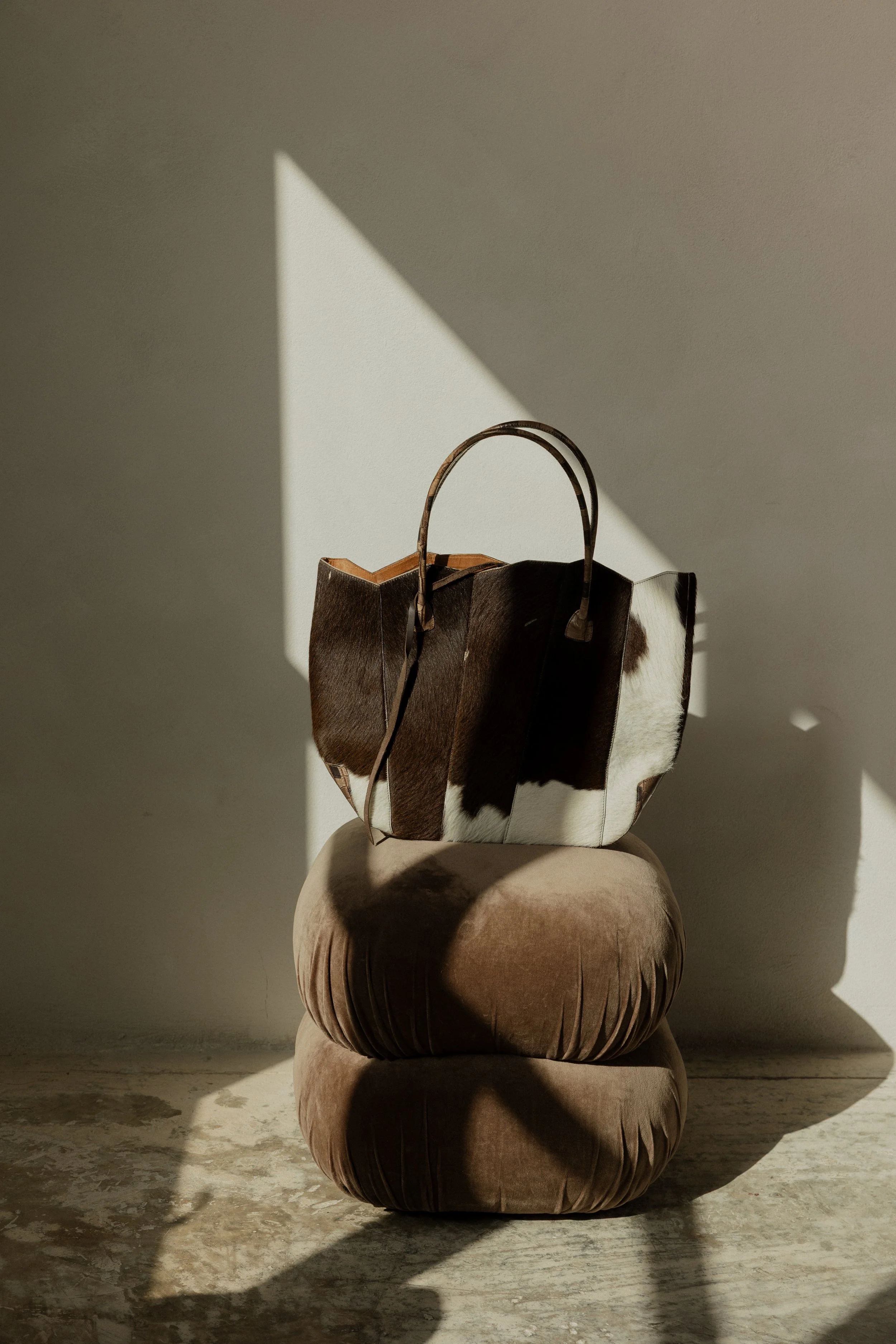 A brown and white cowhide-patterned bag resting on a beige cushioned ottoman, sunlight casting shadows on a plain wall and concrete floor.