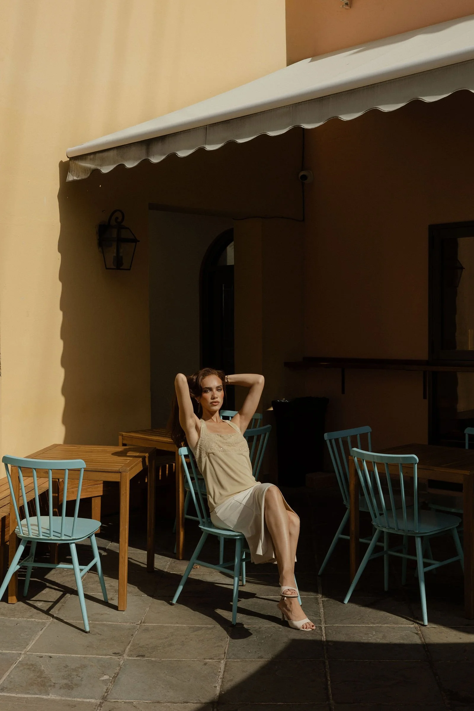 A young woman with brown hair and light skin is sitting on a blue chair outdoors, wearing a beige dress and white high-heeled sandals, with her arms raised behind her head. She is surrounded by wooden tables and blue chairs, with a yellow wall, an awning, a lantern, and a window in the background, under clear daylight.