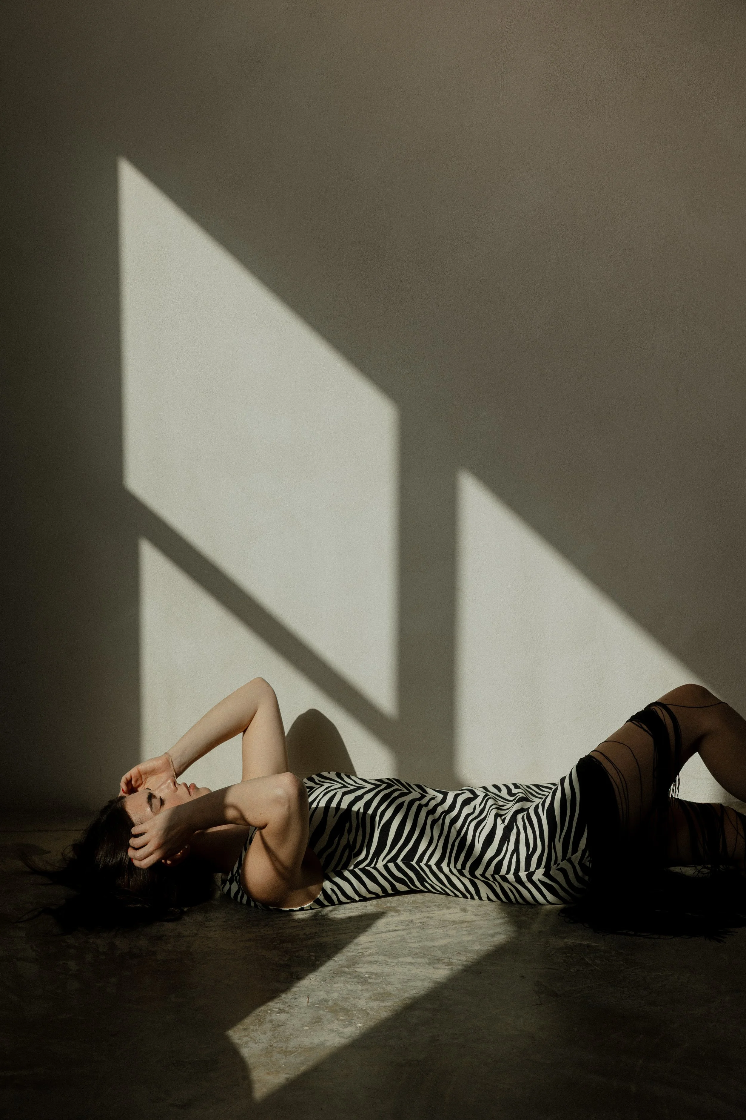 A woman lying on the floor in a zebra print dress, with dark hair spread out, striking a relaxed pose with one arm over her head, surrounded by dramatic shadows cast by sunlight through a window.