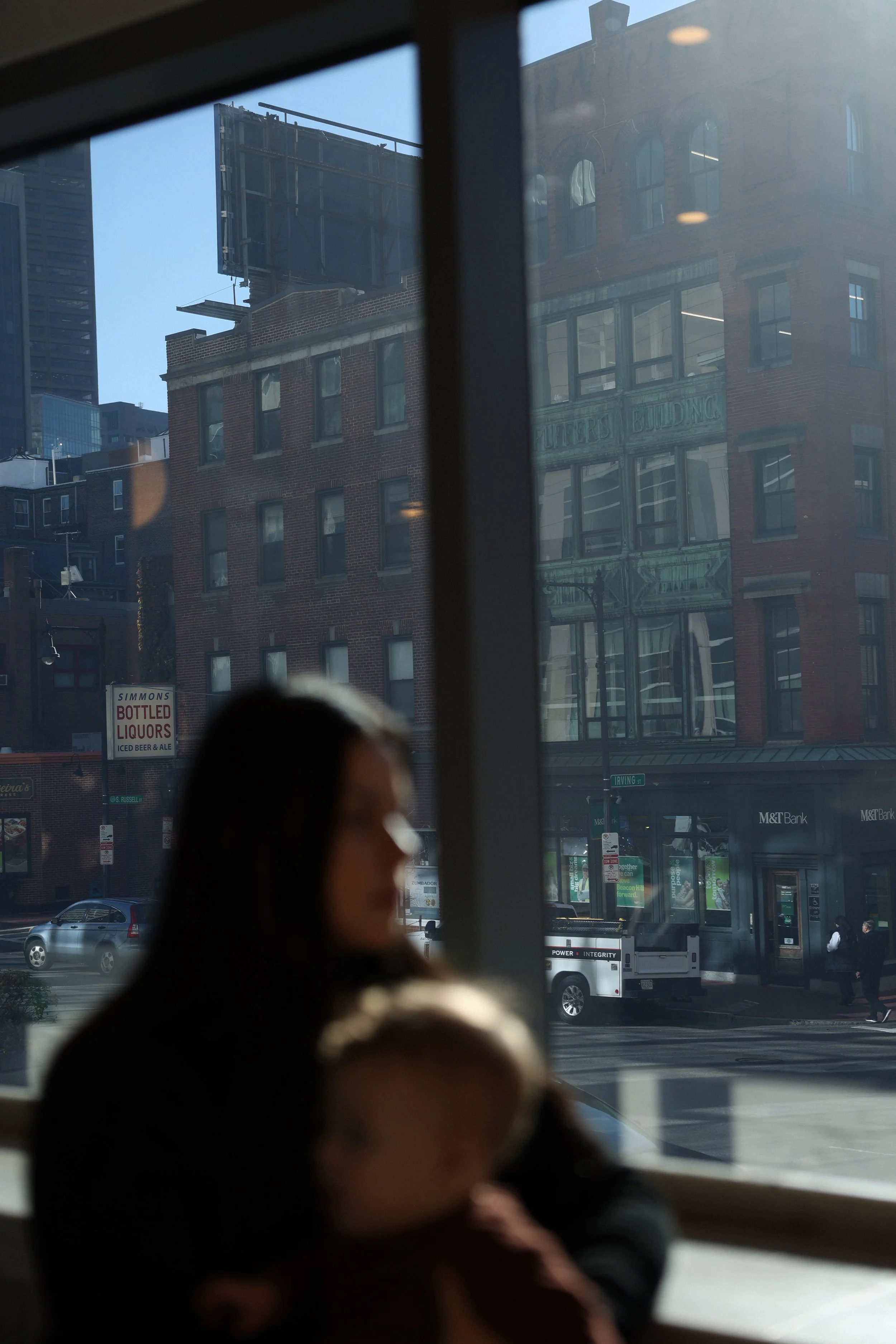 A city street scene viewed through a window with white frames. The street has parked cars and people walking. Several buildings are visible, including one with a sign for bottled liquors, and another with a glass entrance and the words 'BUILDINGS' embossed on it. There is a bus in the street with the words 'POWER' and 'INTEGRITY' on its side.