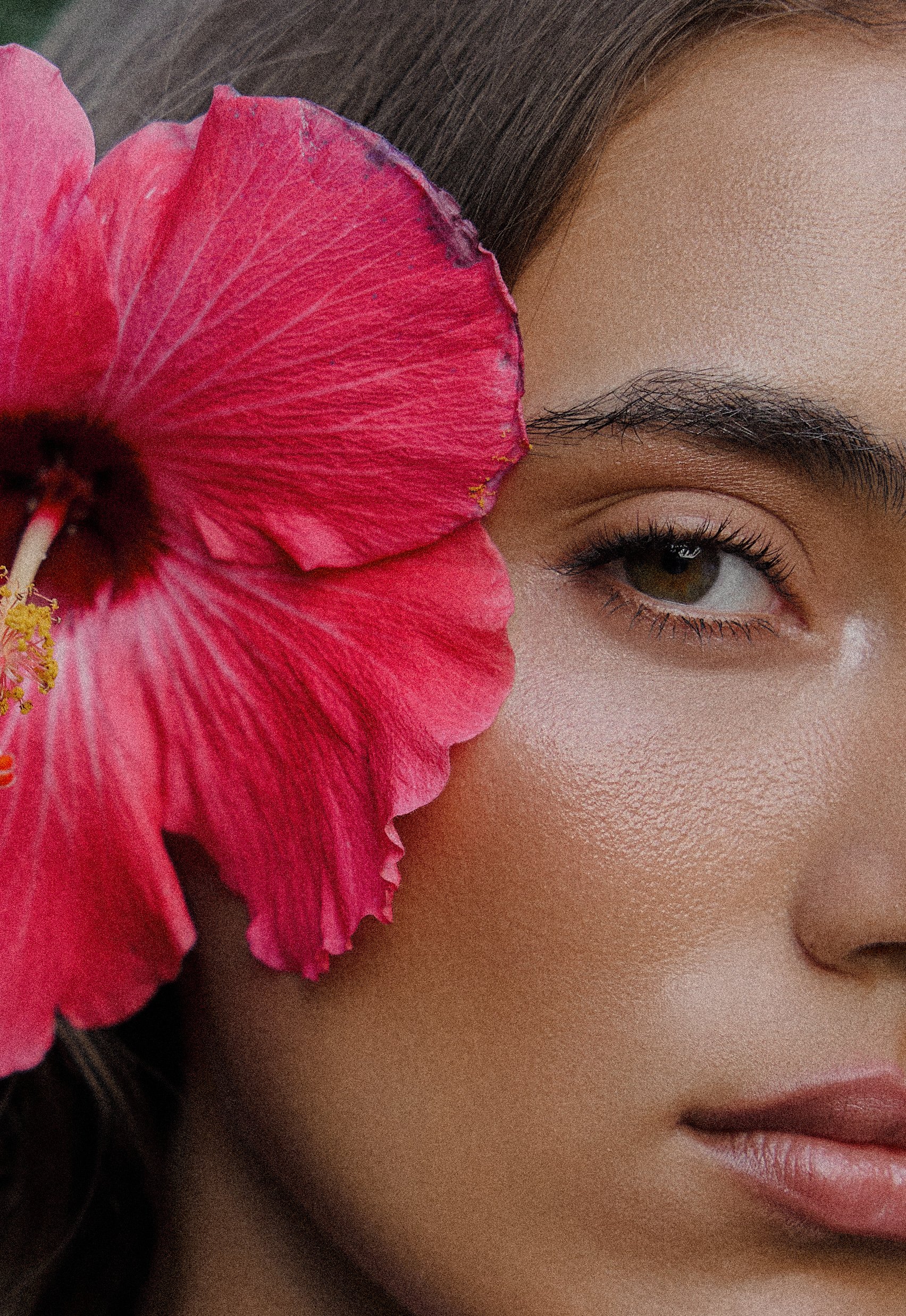 Close-up of a woman's face with a large pink flower covering one eye, showing her other eye, eyebrow, and part of her lips.