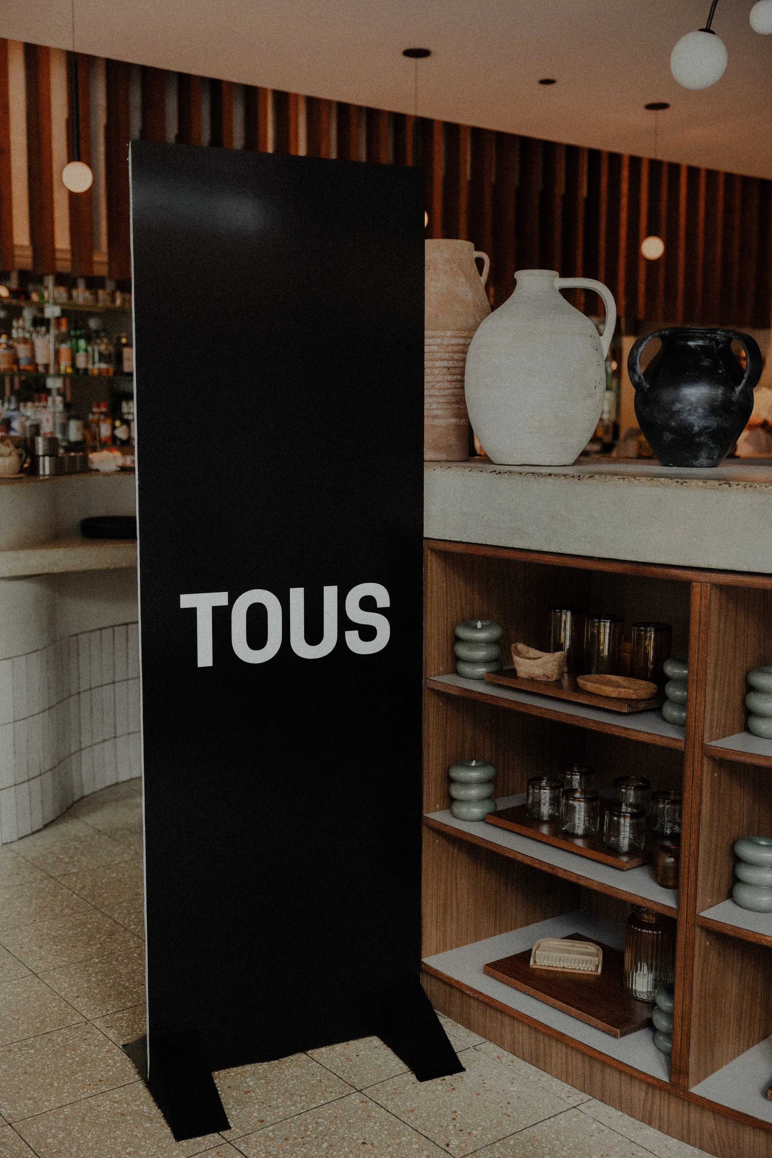 Display shelf with pottery and glassware, partially obscured by a black sign with white text, in a modern interior with wood paneling and round pendant lights
