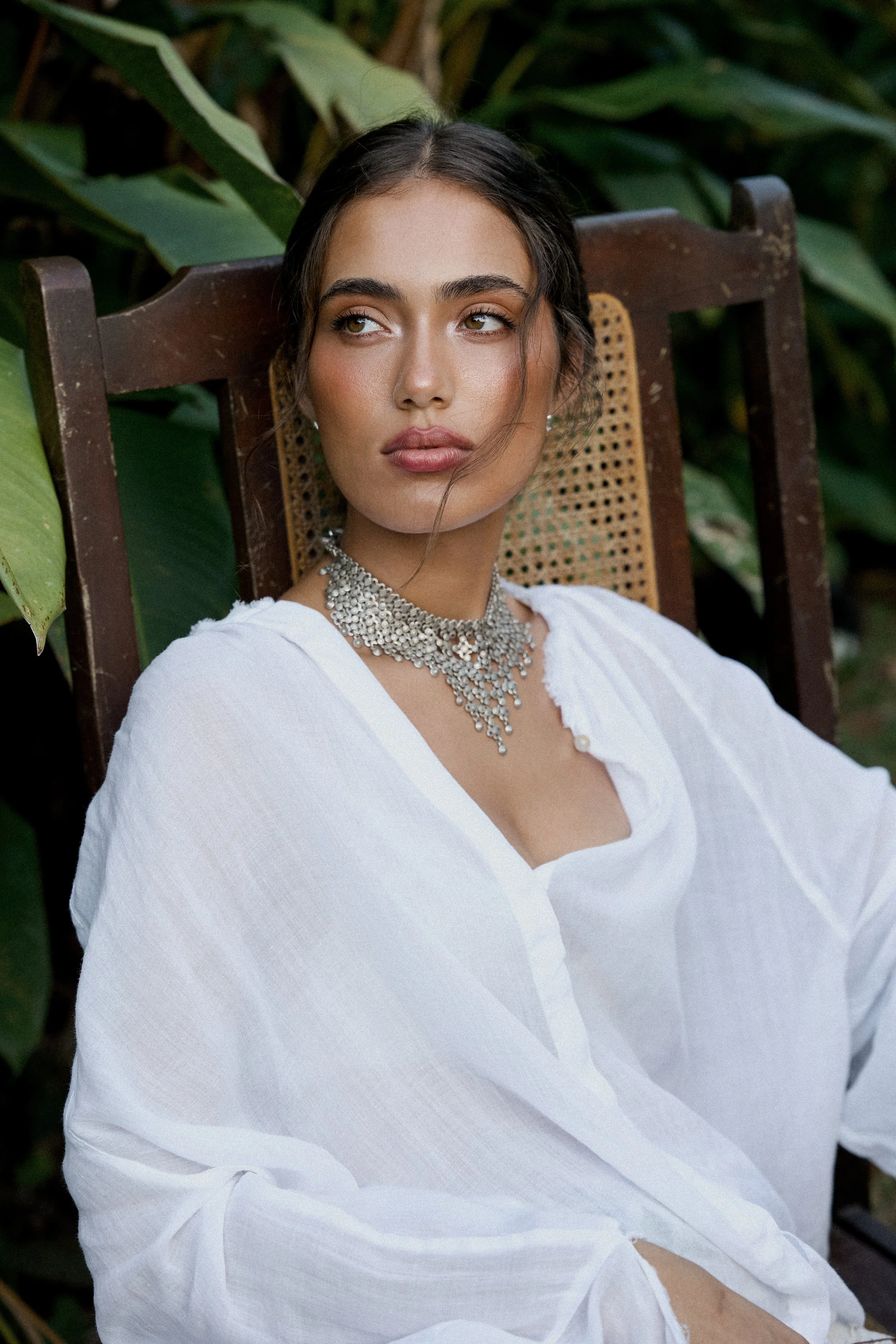 A woman sitting outdoors on a brown wooden chair with a rattan backrest, surrounded by green leafy plants, wearing a white outfit and a sparkling silver necklace, looking to her left with a neutral expression.