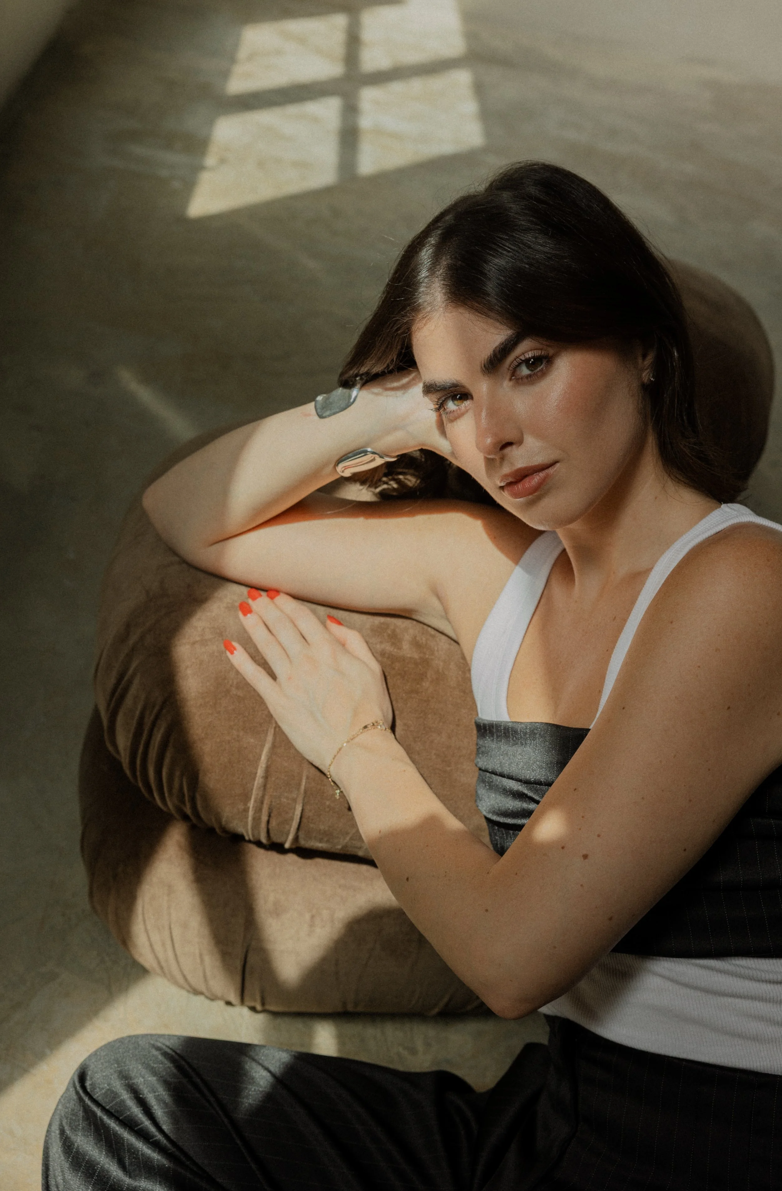 A woman with dark hair and light skin sitting on a beige couch, looking at the camera, with sunlight casting a shadow of a window on the wall behind her.