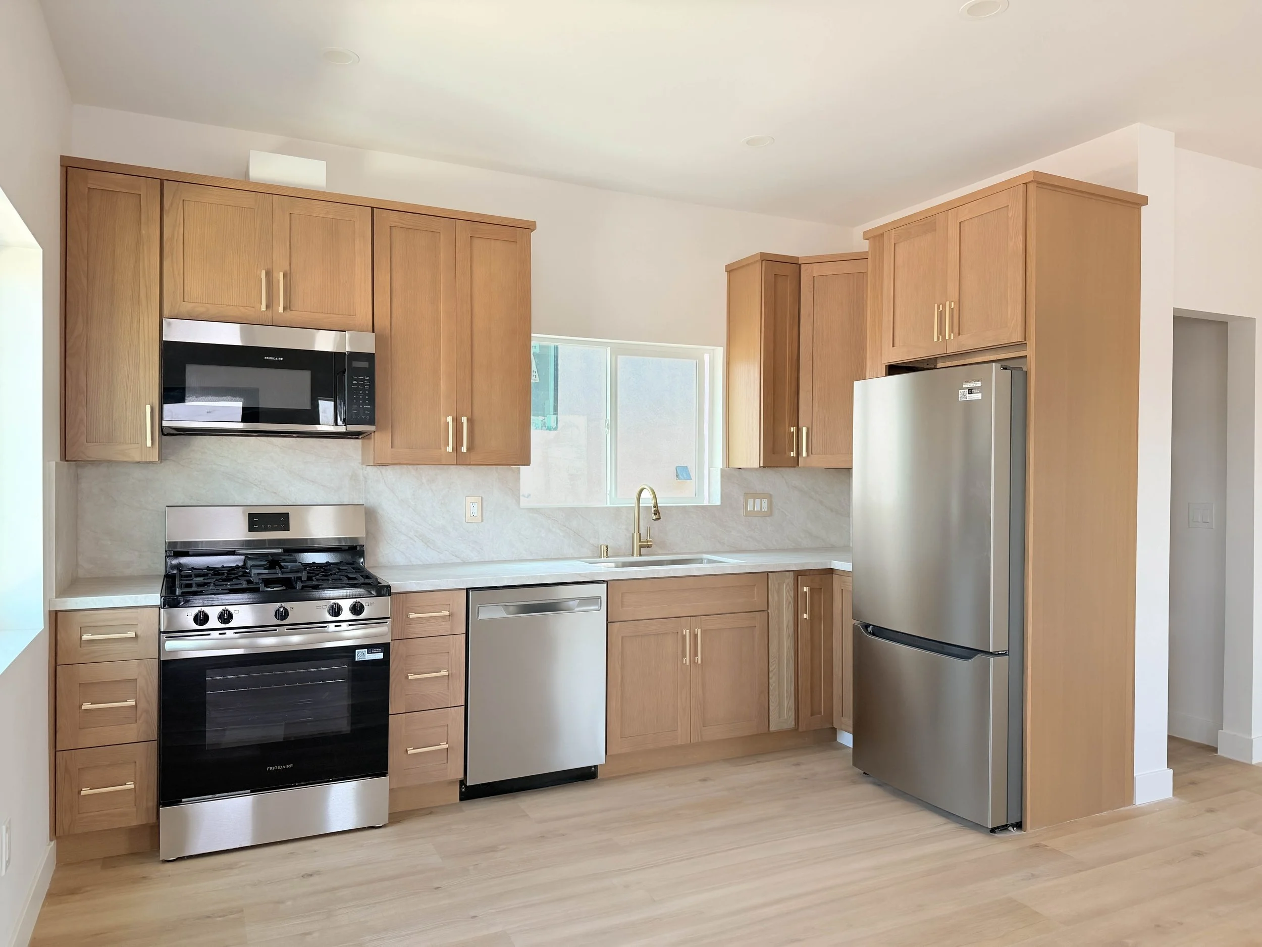 Modern kitchen with wooden cabinets, stainless steel microwave, oven, and refrigerator, light-colored walls, and hardwood floor.
