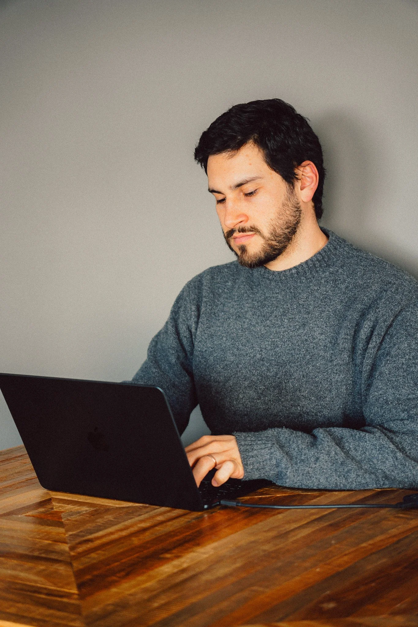 A man with dark hair and a beard sitting at a wooden table, working on a black laptop, wearing a gray sweater, against a plain gray wall.