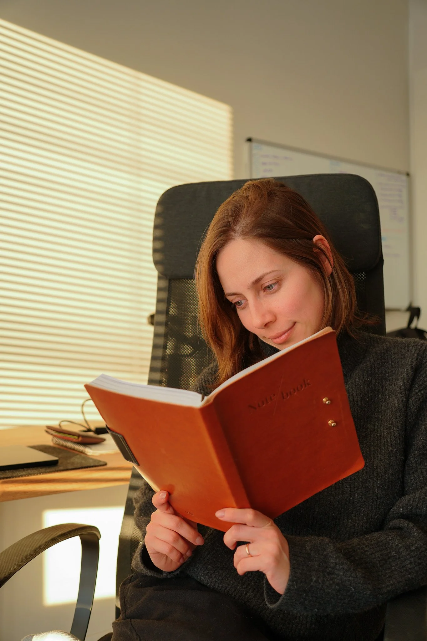 A woman sitting in an office chair, reading a notebook or journal, with a desk and window blinds in the background.