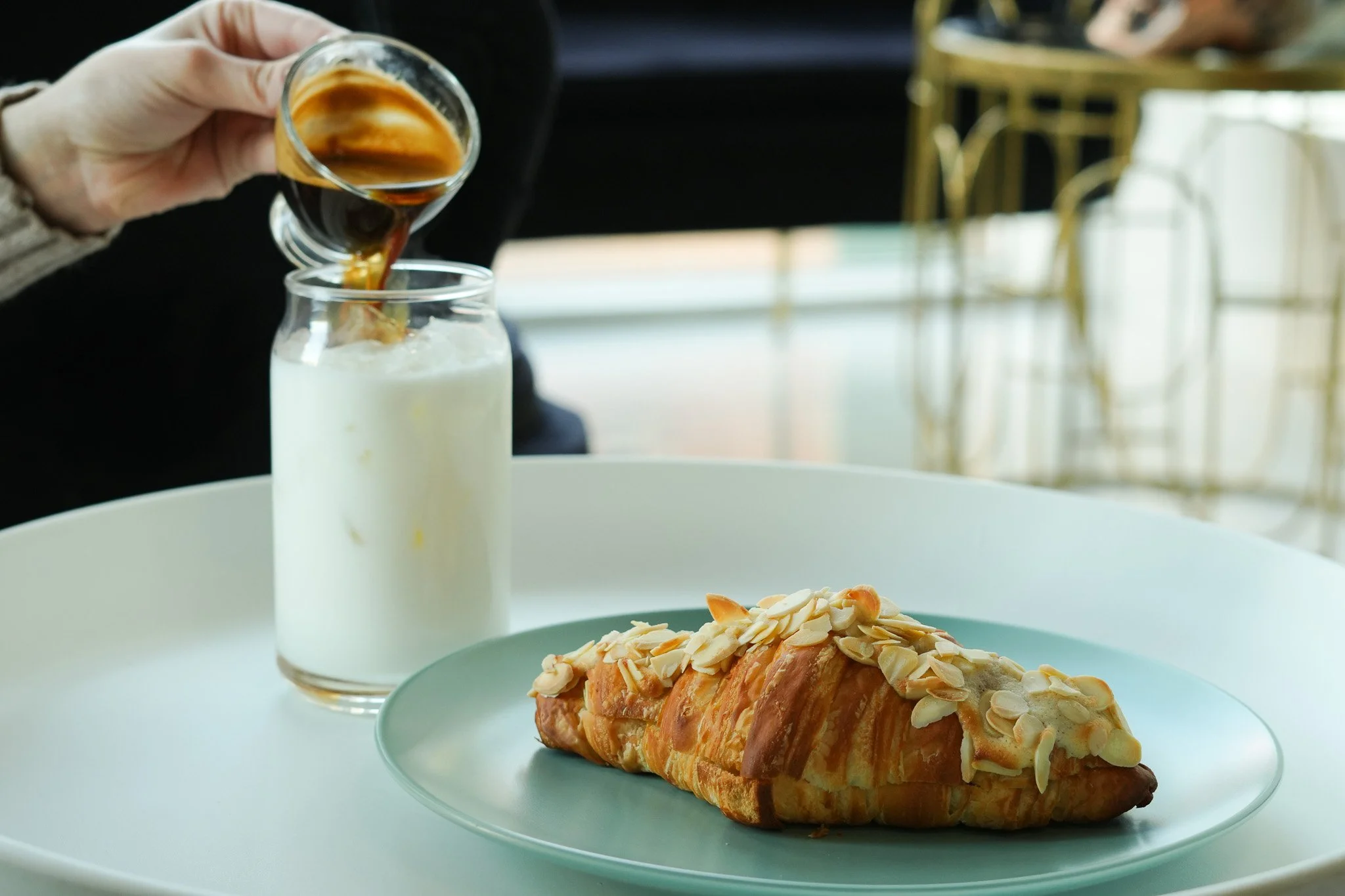 A croissant topped with sliced almonds on a green plate, with coffee being poured into a glass of milk on a white tray.