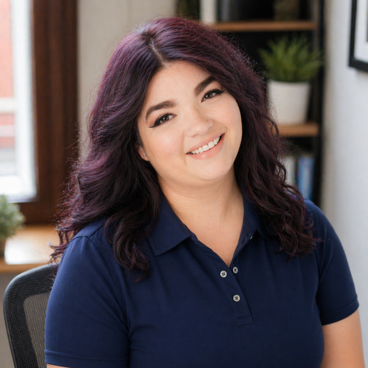 A woman with long, dark red hair smiling and sitting in a room with bookshelves and houseplants in the background.