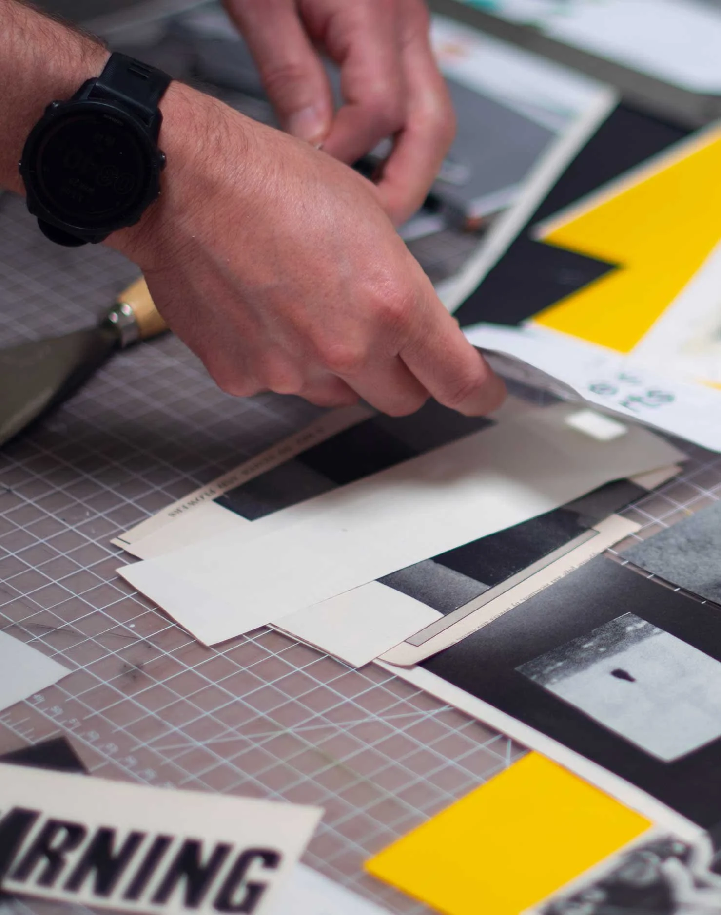 Close-up of a person's hand flipping through printed color swatch sheets or labels, with a black watch on their wrist, on a grid cutting mat.