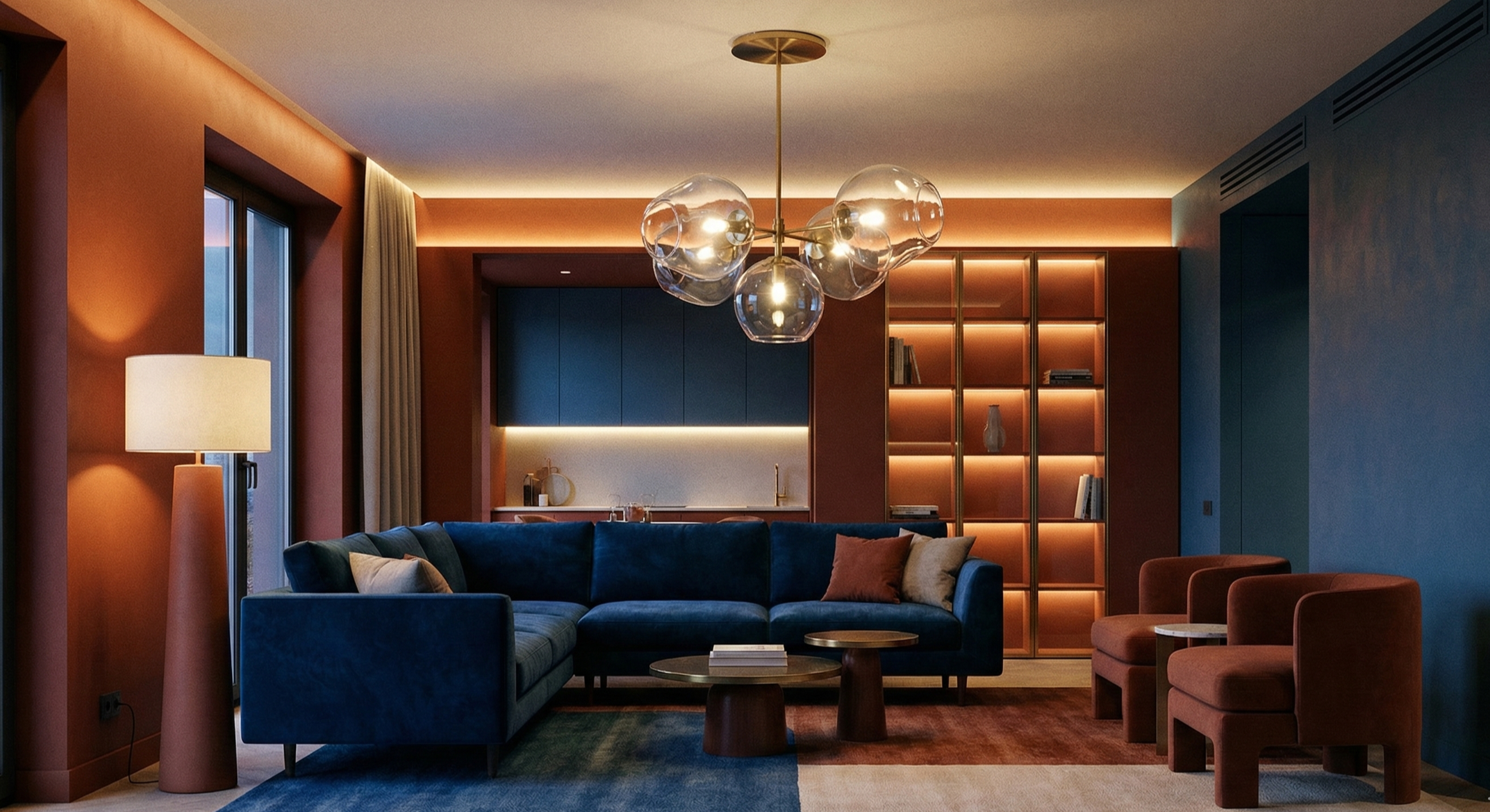 Modern living room with a large blue sectional sofa, two rust-colored armchairs, round wooden coffee tables, a floor lamp near a window, and a ceiling light fixture with glass globes. The back wall features built-in shelves with lighting and a small open kitchen area with dark cabinets and under-cabinet lighting.