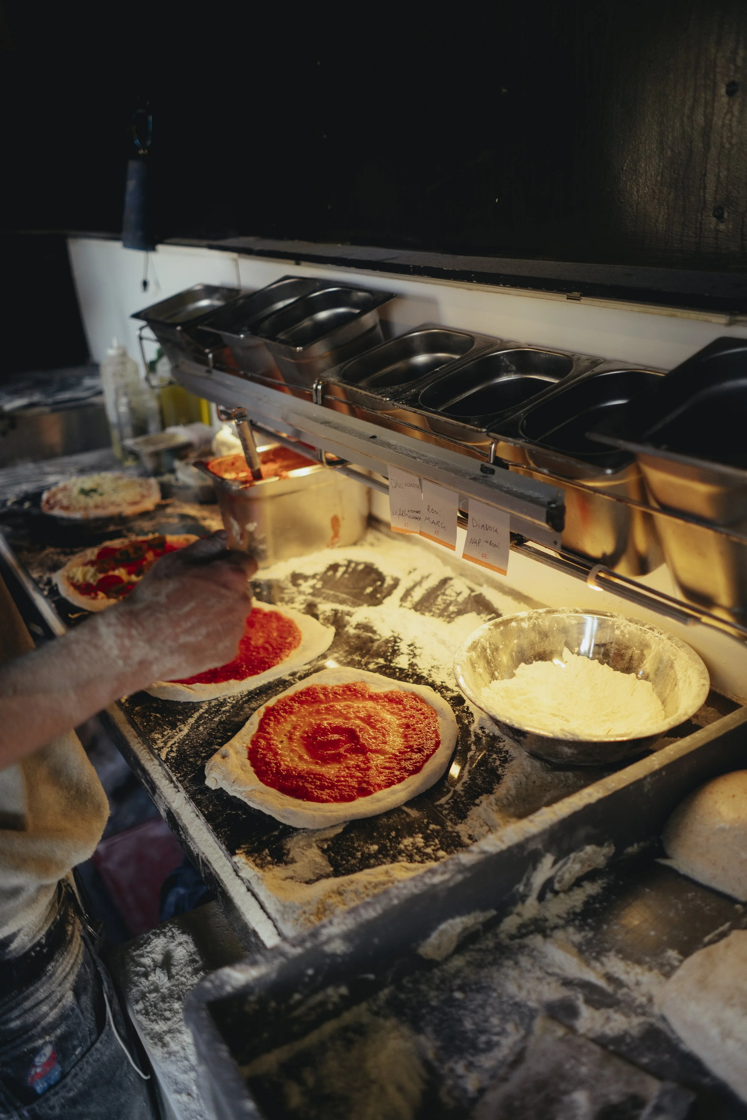 A person preparing pizza dough with tomato sauce on a floured work surface in a kitchen, with pizza toppings and ingredients nearby.