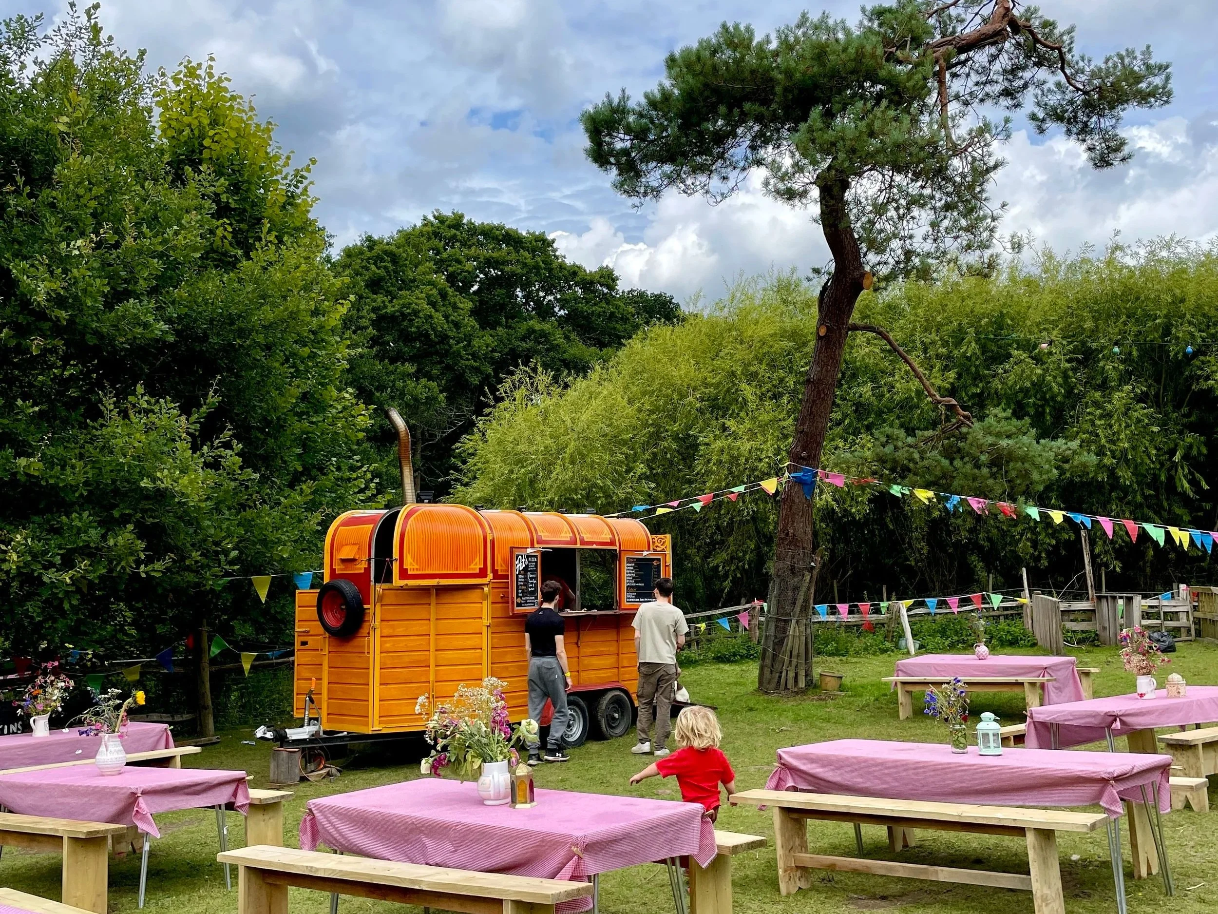 A colorful outdoor food truck with two people in front, set in a garden with wooden picnic tables covered in pink tablecloths, and decorated with flower vases and lanterns, under a cloudy sky and surrounded by green trees.