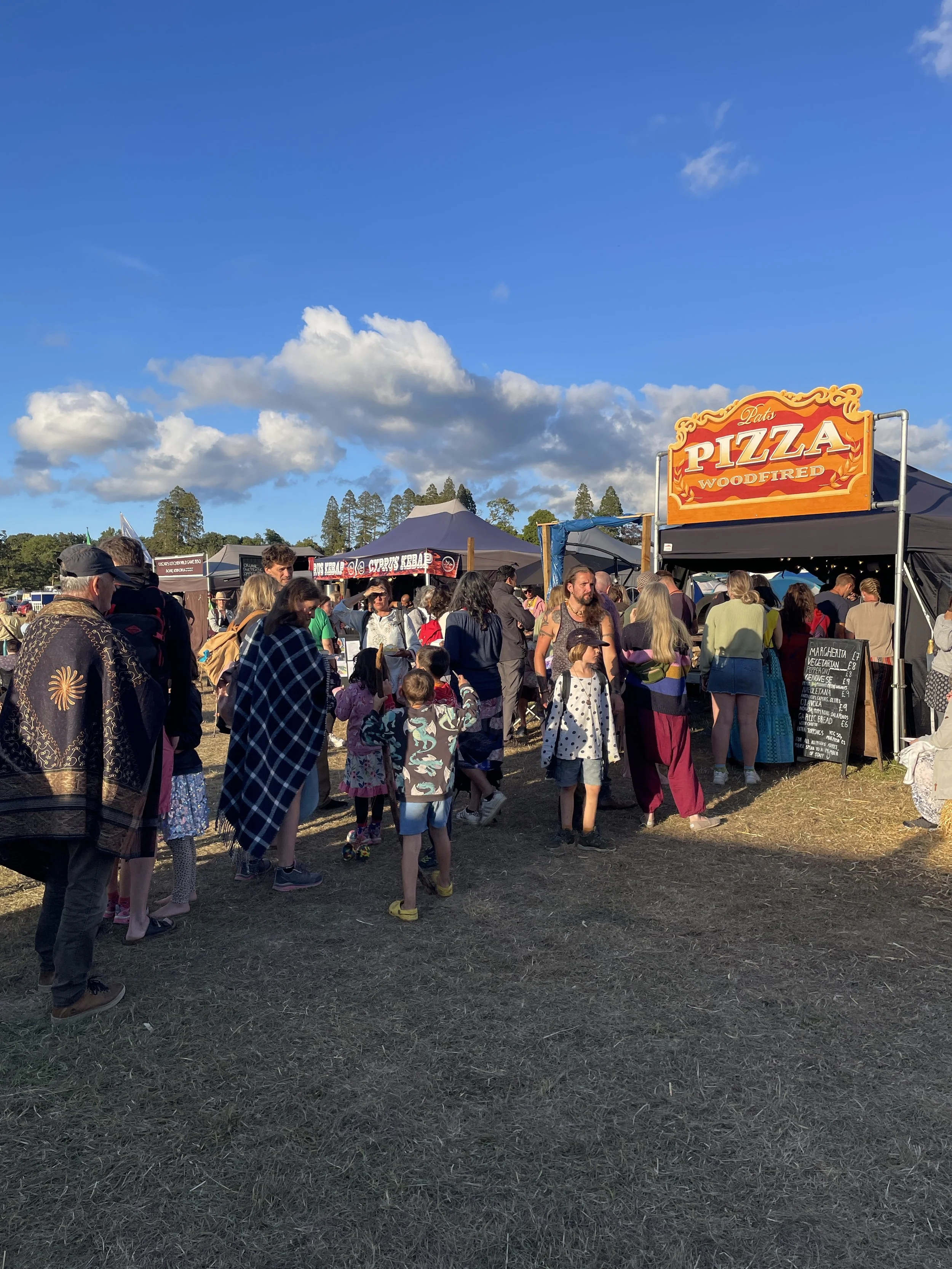 A big queue of people waiting in line at Into the Wild festival, for our pizzas from our large festival Pat's wood-fired Pizza set-up. The sun is shining and the sky is blue with a few puffy clouds.