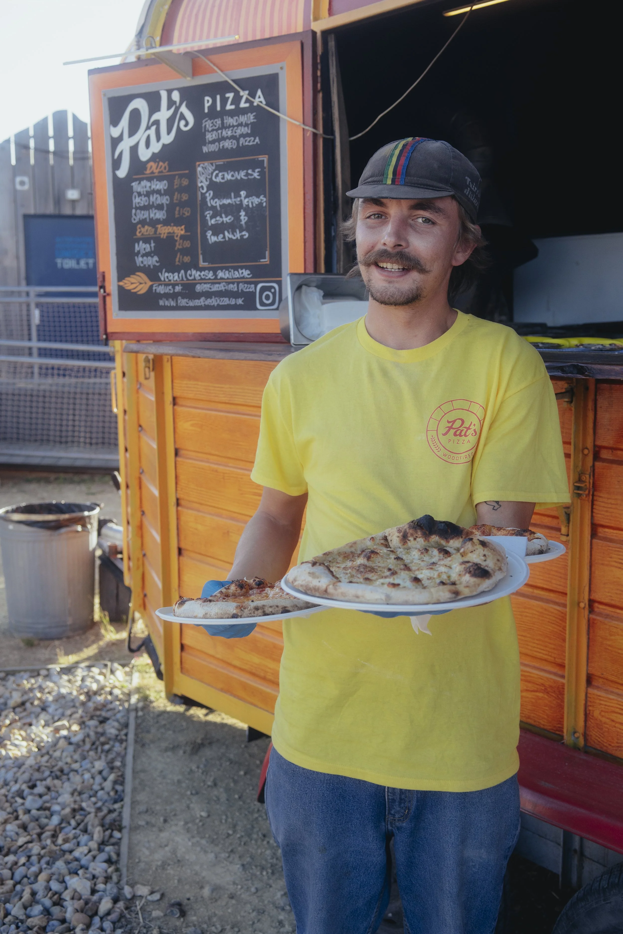 Man with a moustache wearing a yellow Pat's pizza T-shirt and a cycling cap holding three pizzas in front of our funky horsebox trailer that we use for festivals and events.