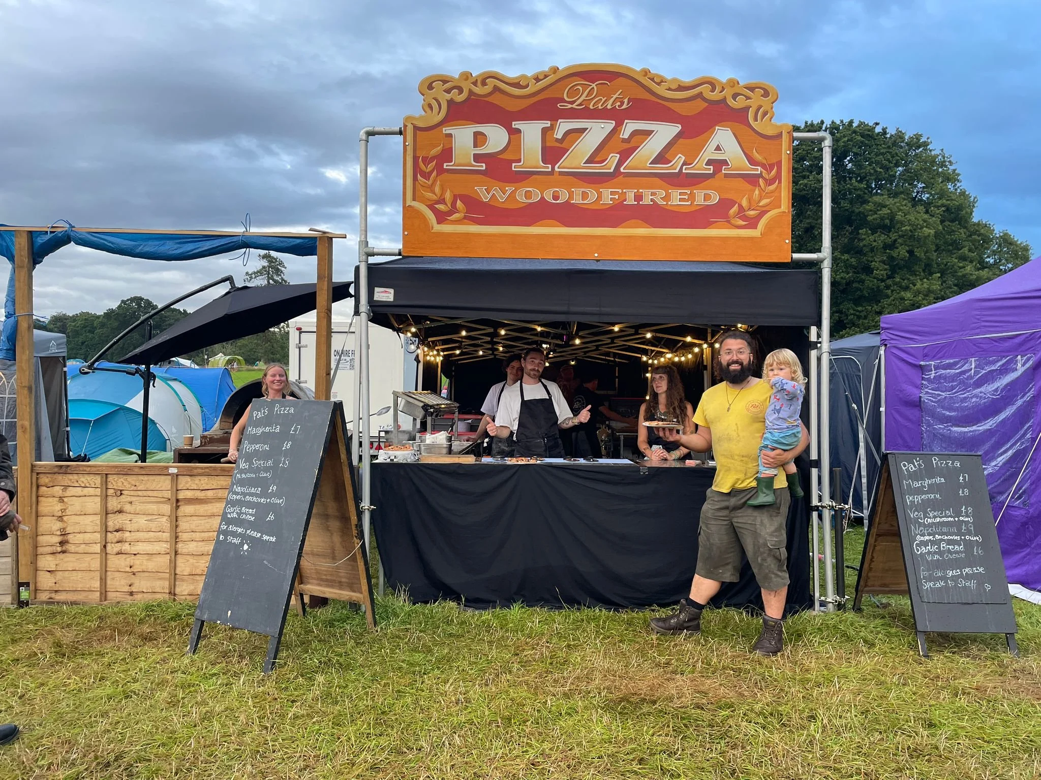 A food stall named 'Pat's Pizza', specializing in wood-fired pizza, set up at an outdoor event, with a man holding a child in front, and staff preparing food inside the stall.
