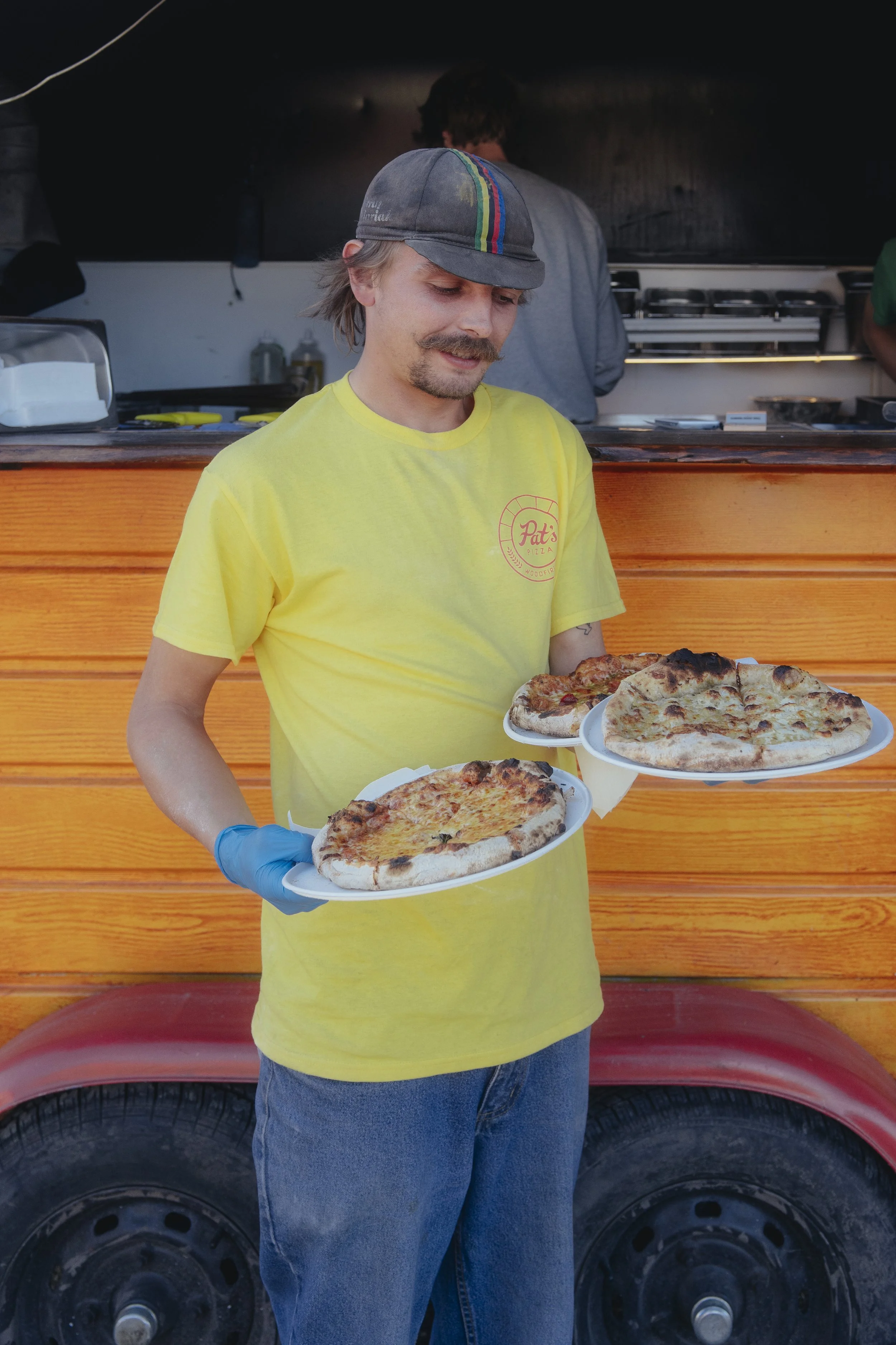 A man wearing a yellow T-shirt, gray cap with colorful stripes, and blue gloves is holding two plates of pizza, with a second man working in the background at a food truck.