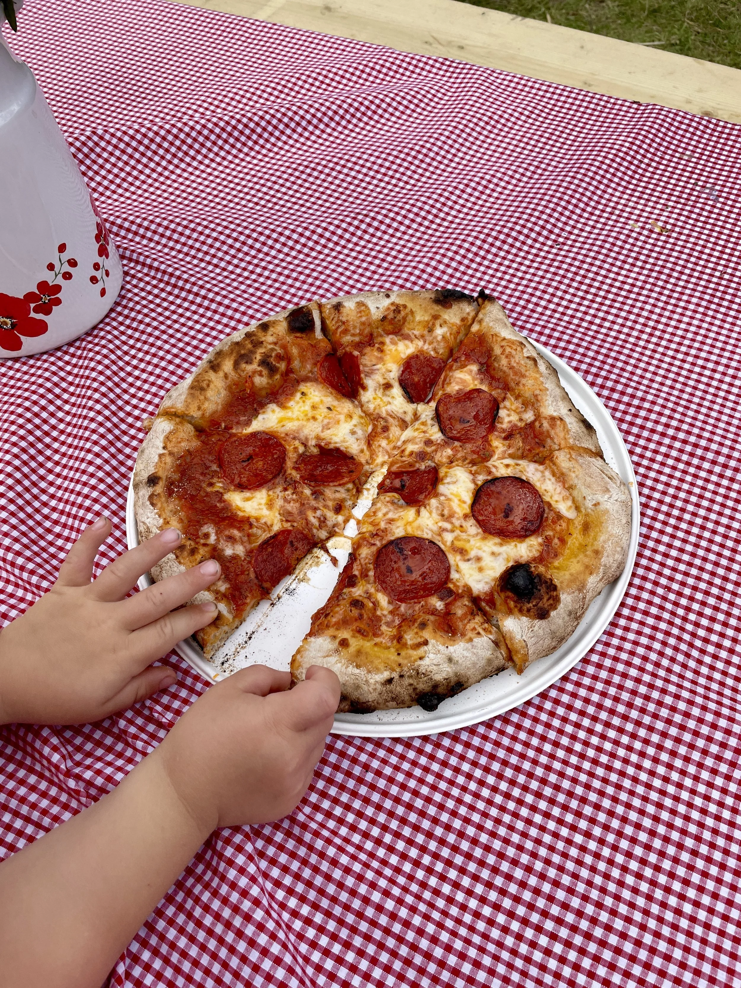 Slice of pepperoni pizza on a white plate placed on a red and white checkered tablecloth outside; child's hands reaching for the pizza.