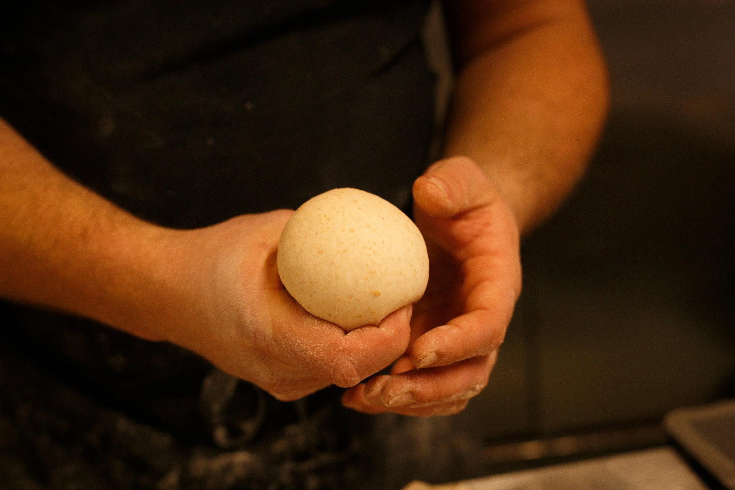 Person holding a round piece of dough, with hands covered in flour, in a kitchen setting.