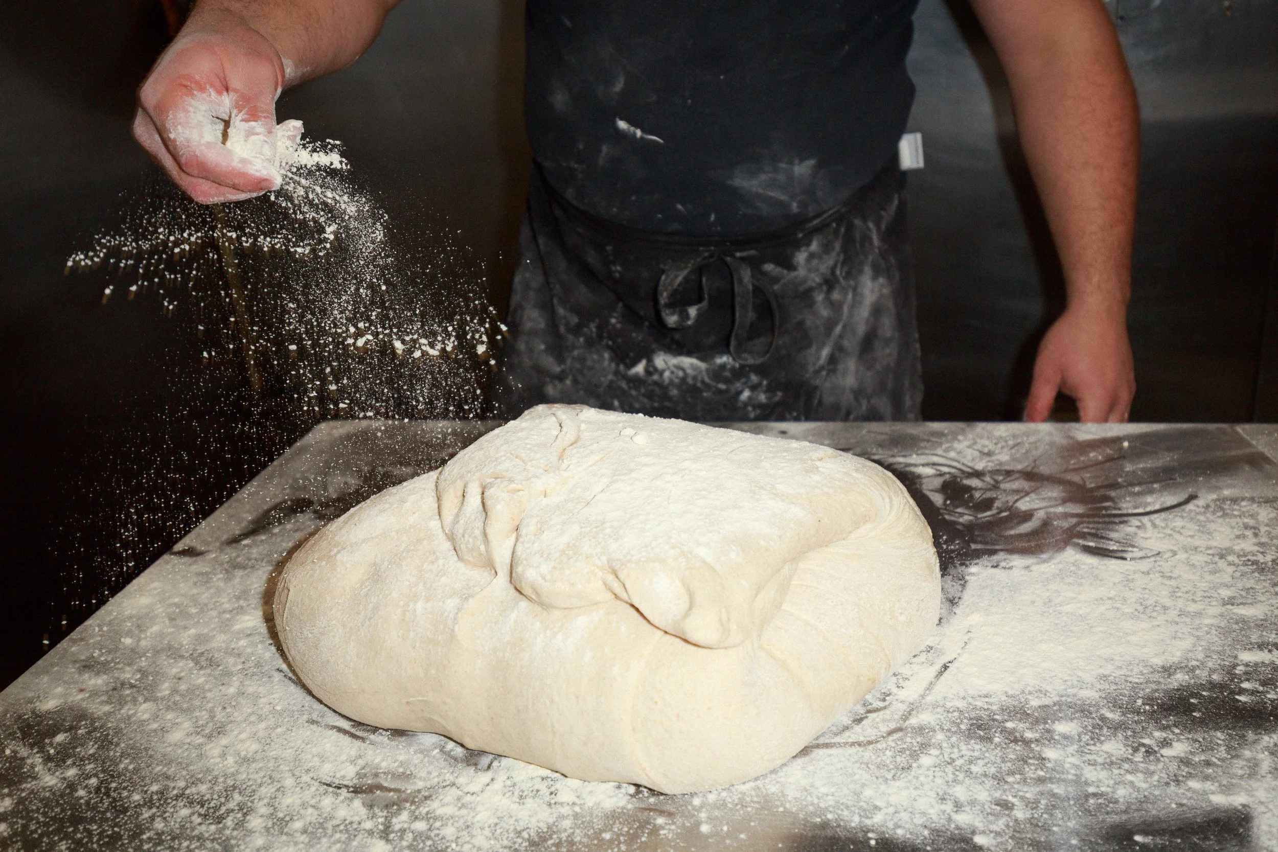 Person sprinkling flour on a mound of dough on a floured surface in a bakery kitchen.