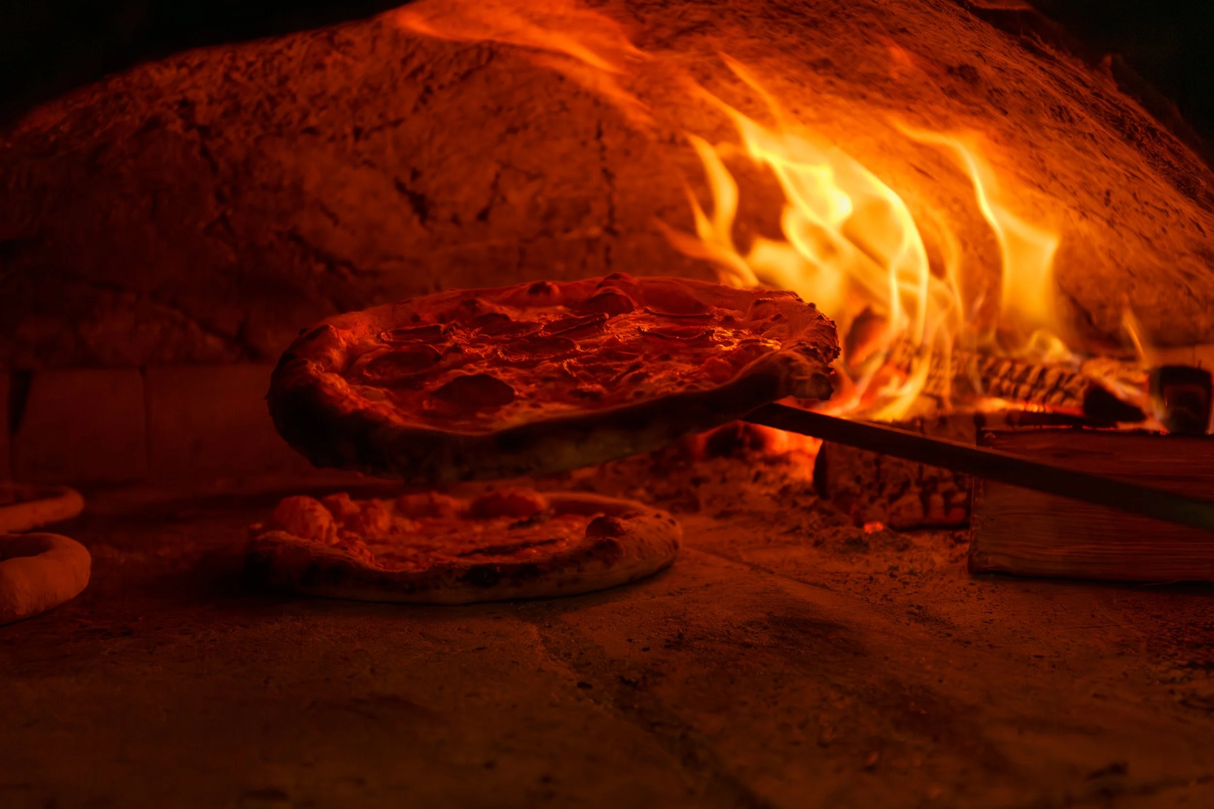 A pepperoni pizza being held up by the pizza peel in our hand-built wood-fired oven, at The Lewes Arms pub, with flames licking around the pizza.