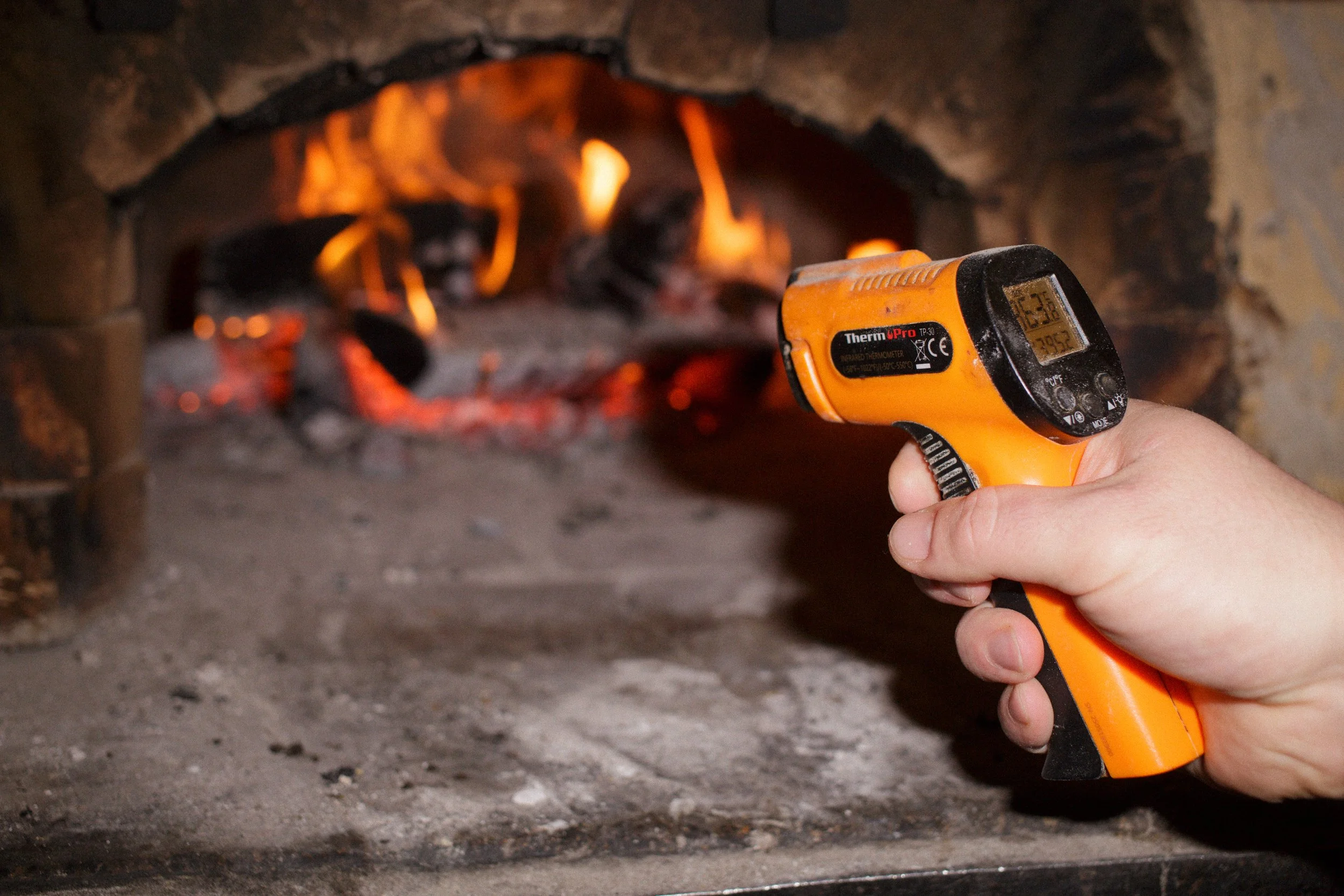 Pat is holding a temperature gun to measure the temperature inside the hand-built wood-fired oven with visible flames and glowing embers.