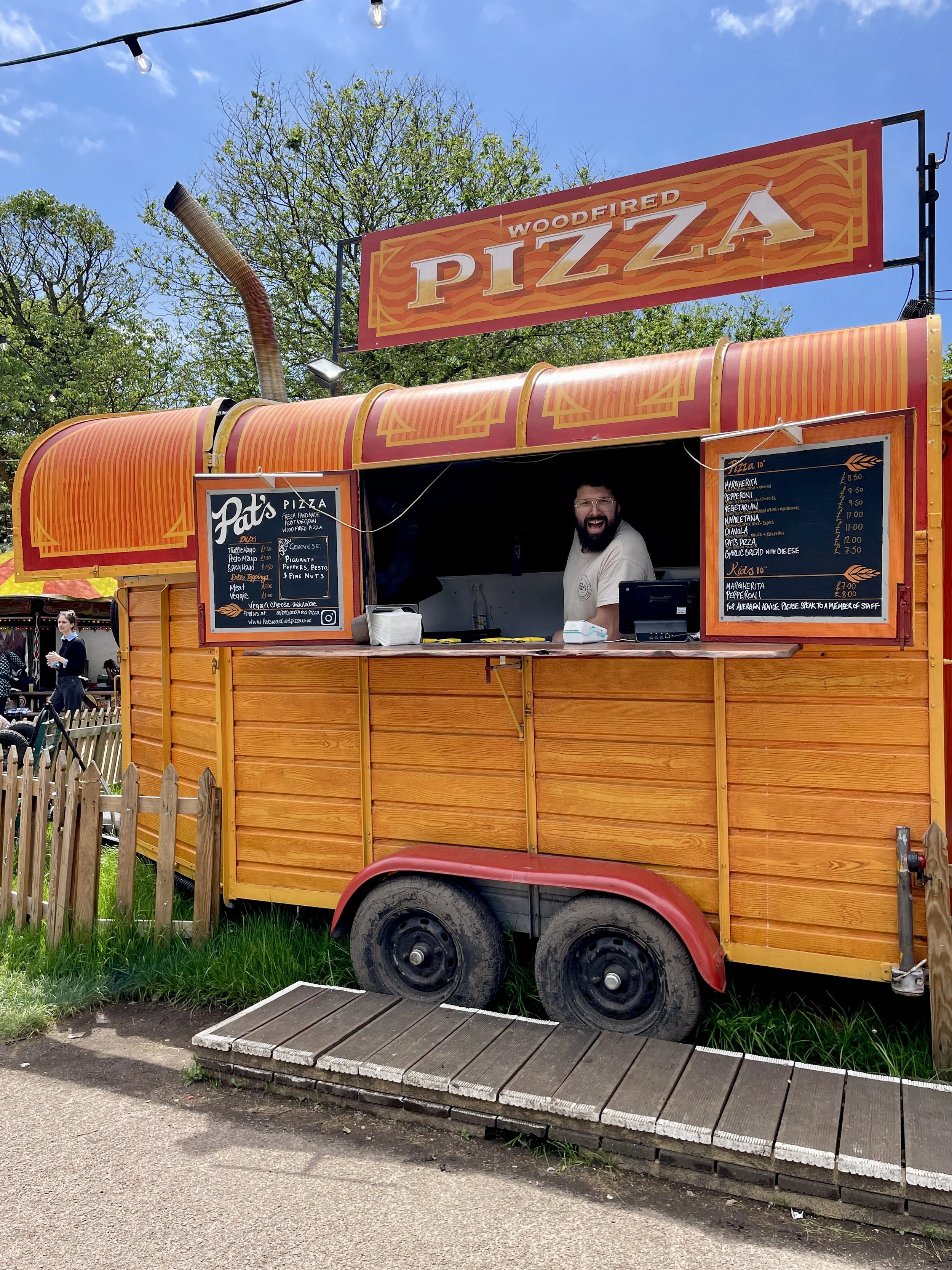 A man working at a bright orange wood-fired pizza food truck with a red and yellow sign reading 'Woodfired Pizza'. The truck has a menu board and is parked next to a wooden walkway.