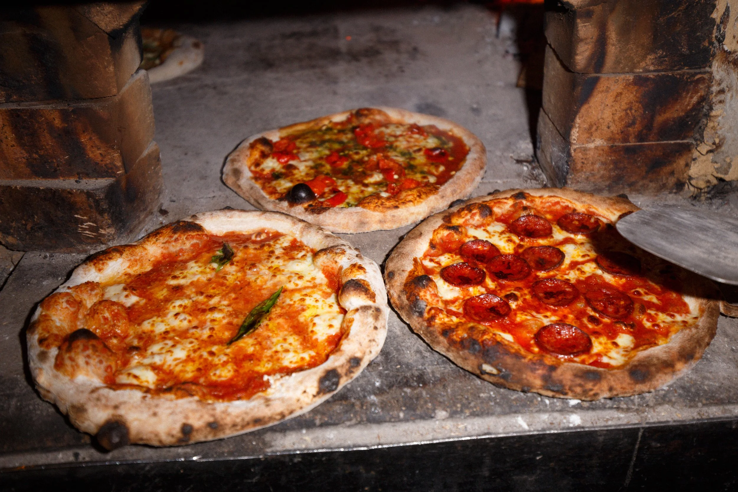 Three pizzas baking in our wood-fired oven, one with pepperoni, another with vegetables, and a Margherita pizza, with a pizza peel visible.