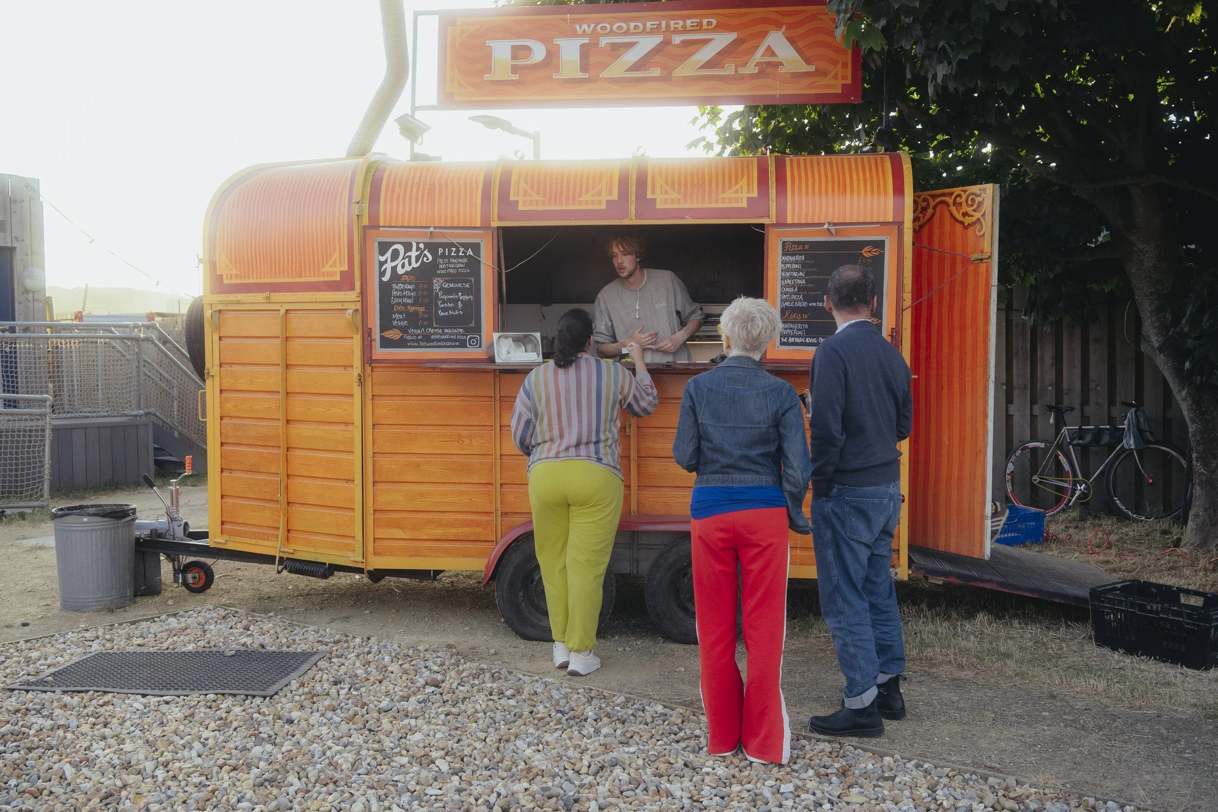 A bright orange wood-fired pizza food truck with a menu board, serving three customers standing in front, including two women and one man, as the person inside the truck prepares food.