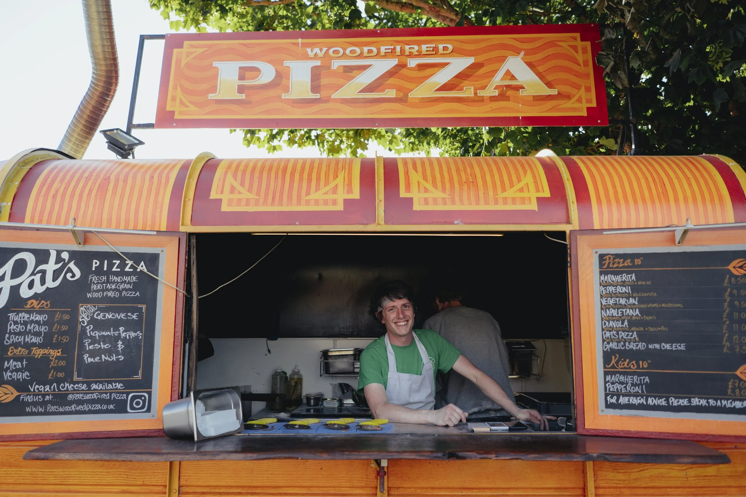 Smiling man in green shirt and apron working at a pizza stand with menu boards displaying various pizza options and toppings, outdoor setting with trees.