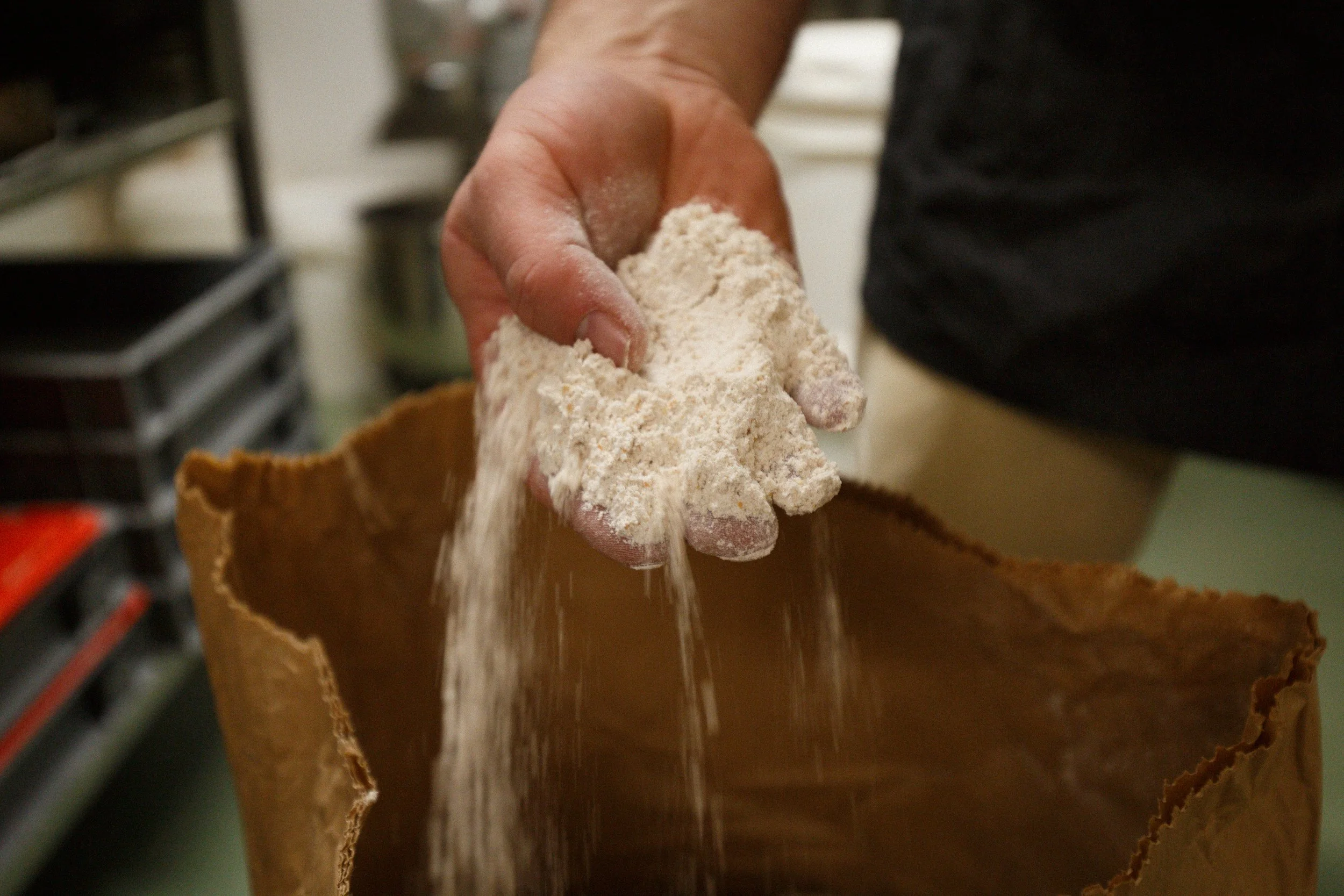 A hand holding white flour, pouring it into a brown paper bag, in a kitchen setting.