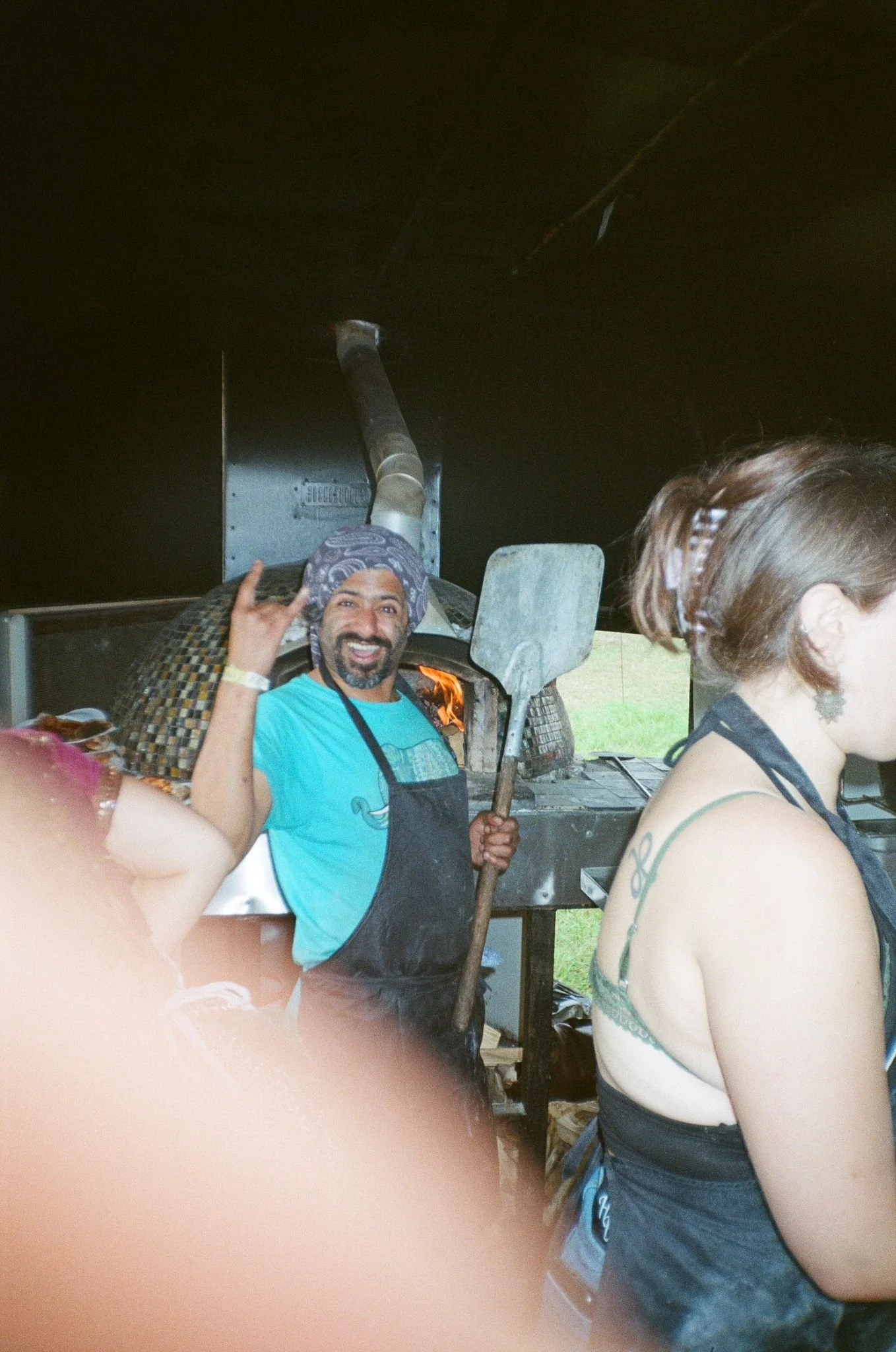 A man with a beard and a bandana, smiling and making a rock sign with his hand, standing in front of a fire inside a pizza oven, holding a pizza peel. A woman with short hair and a tattoo on her shoulder is in the foreground, partially visible, facin
