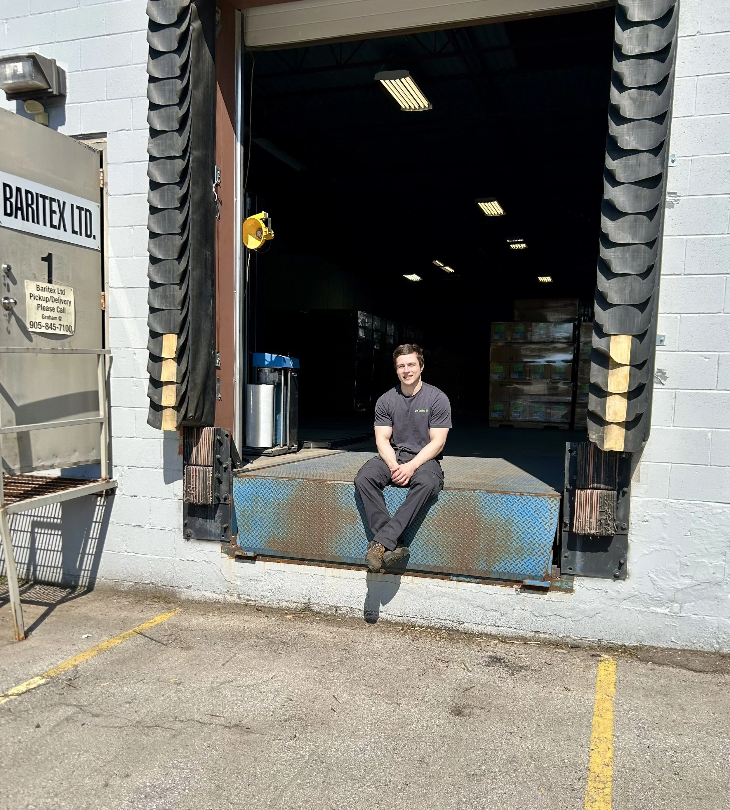 A man sitting in front of a warehouse or industrial building, smiling at the camera. The building has an open loading dock with a black rubber bumper around the door frame, and a small window air conditioning unit on the wall. There is a metal rack outside the building and parking lot lines on the ground.