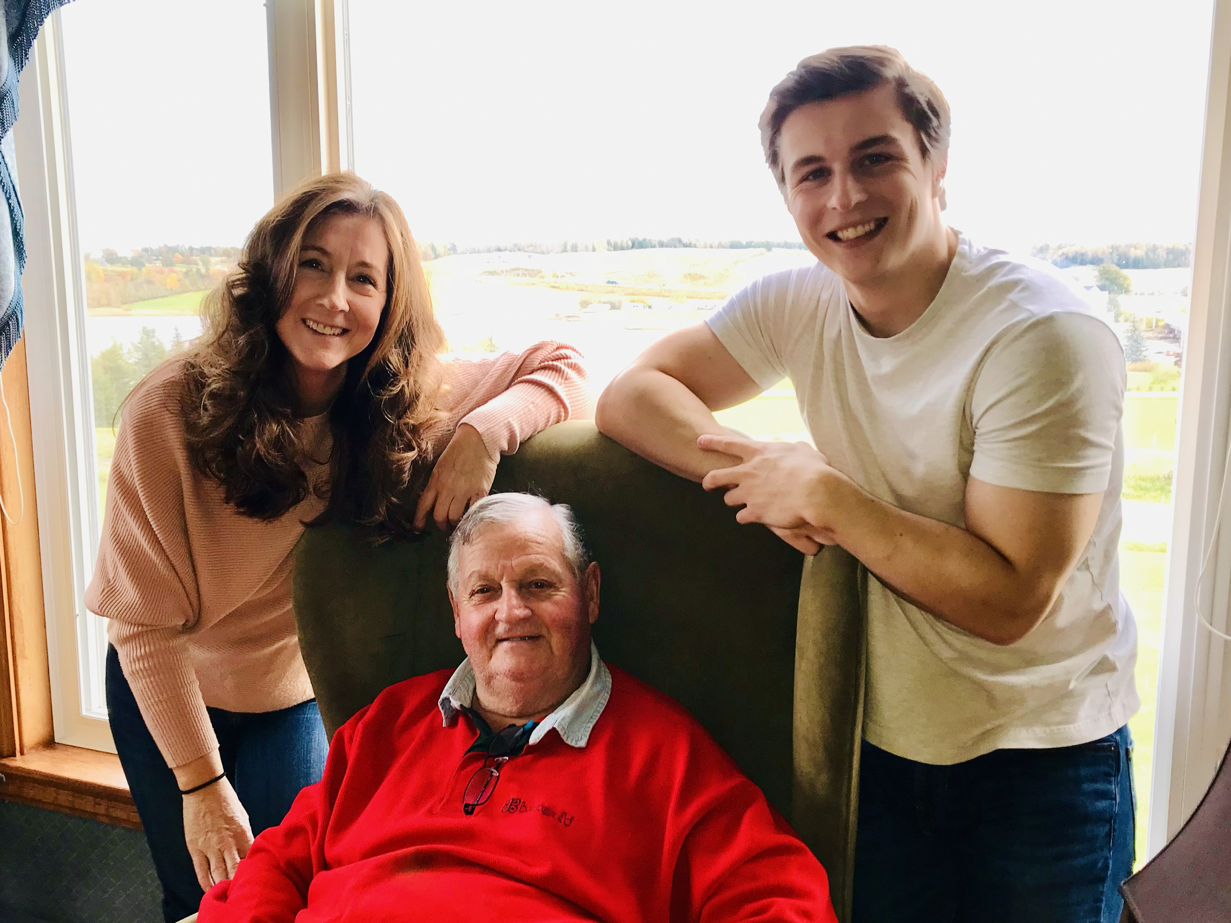 Smiling woman with brown hair, smiling man with dark hair, and an older man in a red sweater, sitting by a window overlooking a rural landscape.