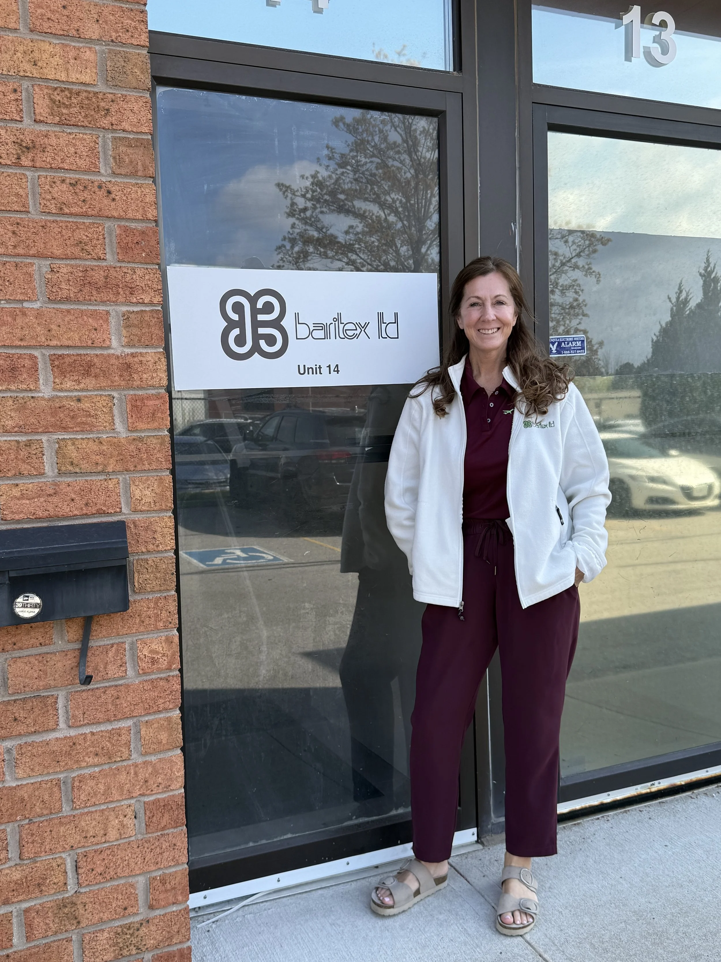 A woman with brown hair and a white jacket standing outside a building with a glass door. The door has a sign that reads '83 Barilex Ltd Unit 14'. The woman is smiling and wearing burgundy pants, a maroon shirt, and beige sandals. There are cars parked in the background.