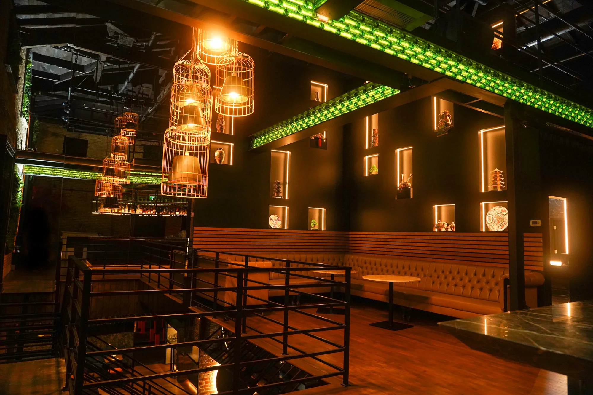 Interior of a modern restaurant or bar with black walls, illuminated display niches with decorative items, hanging orange wire cage pendant lights, and green neon pipe-like lighting fixtures on the ceiling. There's a plush bench seating along the wall and a marble table in the foreground.