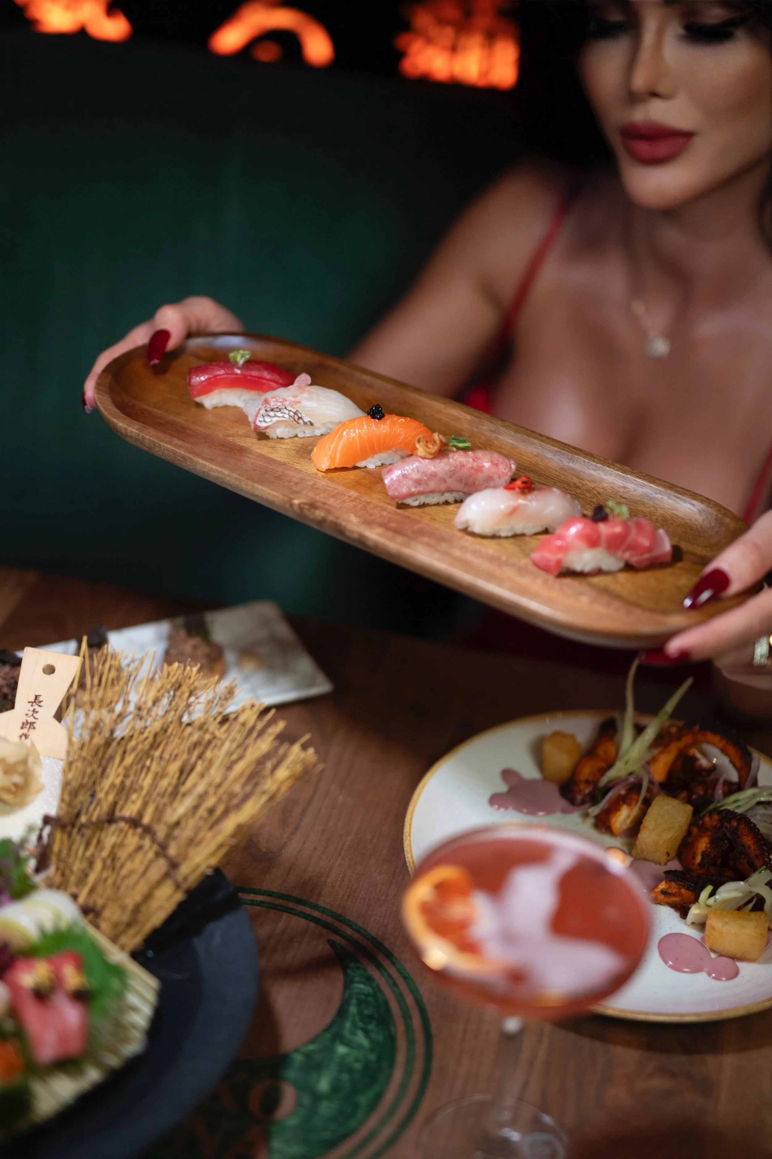 A woman with red lipstick and dark eye makeup holding a wooden platter of assorted sushi, including slices of salmon, tuna, and other fish, at a restaurant table. The table also has other dishes, including a plate of fried food and a glass of pink cocktail.