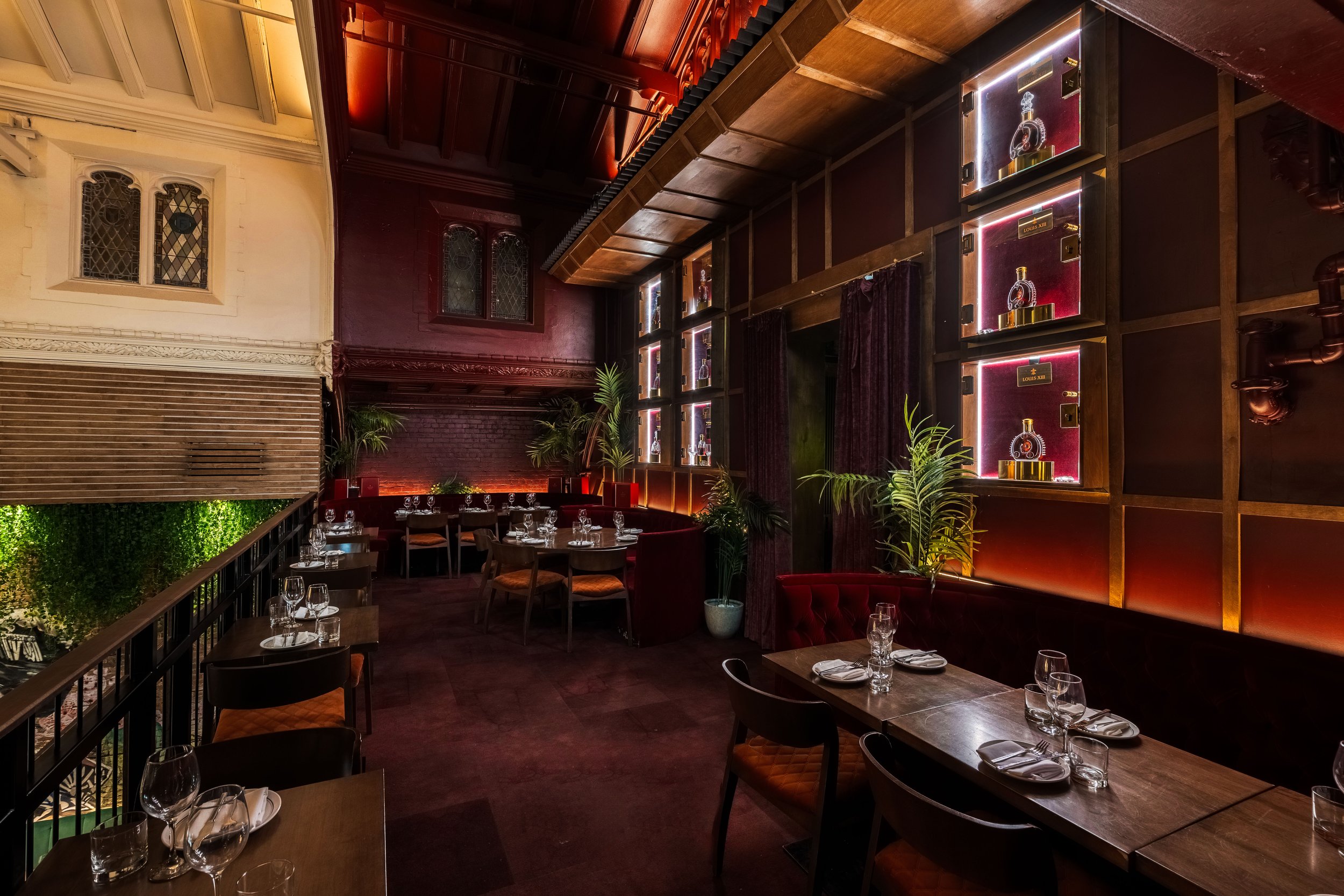Interior of a stylish restaurant with wooden tables set with glasses and silverware, surrounded by upholstered chairs. The walls feature stained glass windows and decorative woodwork, with lush green plants adding a touch of nature.