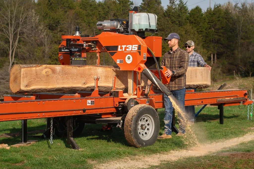 Two men operate a large orange portable sawmill cutting a log into lumber outdoors.