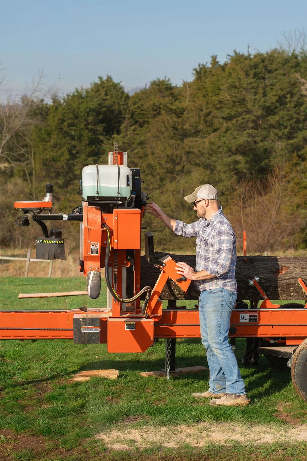 A man operating a portable sawmill outdoors, cutting logs into lumber on a grassy field with trees and a hill in the background.