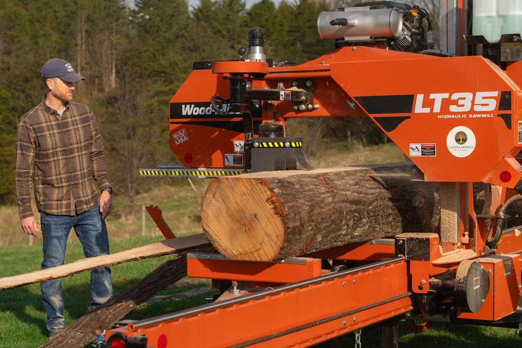 A man wearing a plaid shirt, jeans, and a cap standing next to an orange hydraulic sawmill cutting a large log into smaller pieces outdoors.