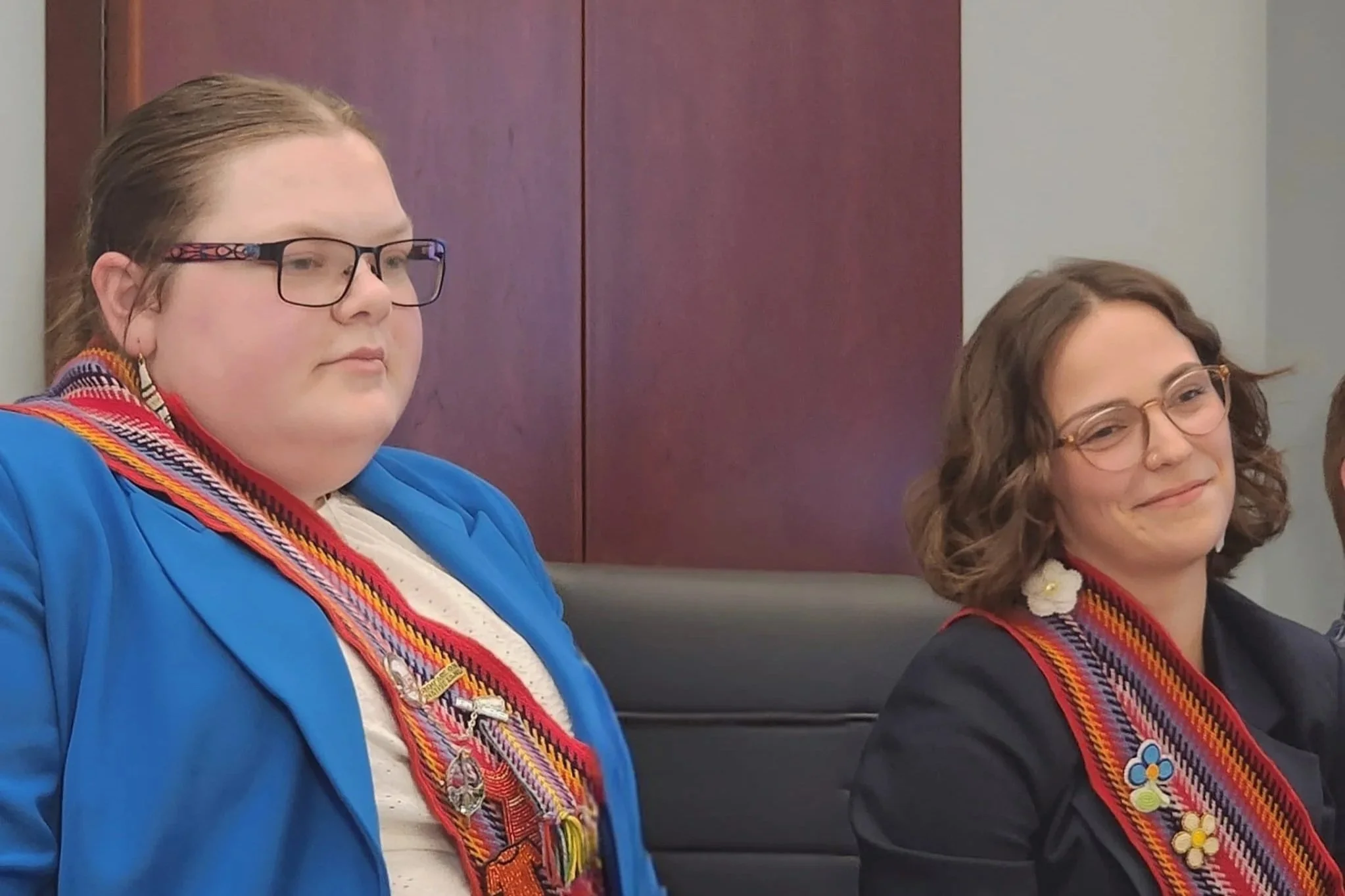 Two young women sitting side by side, wearing formal attire and girl scout sashes with badges, in a professional indoor setting.