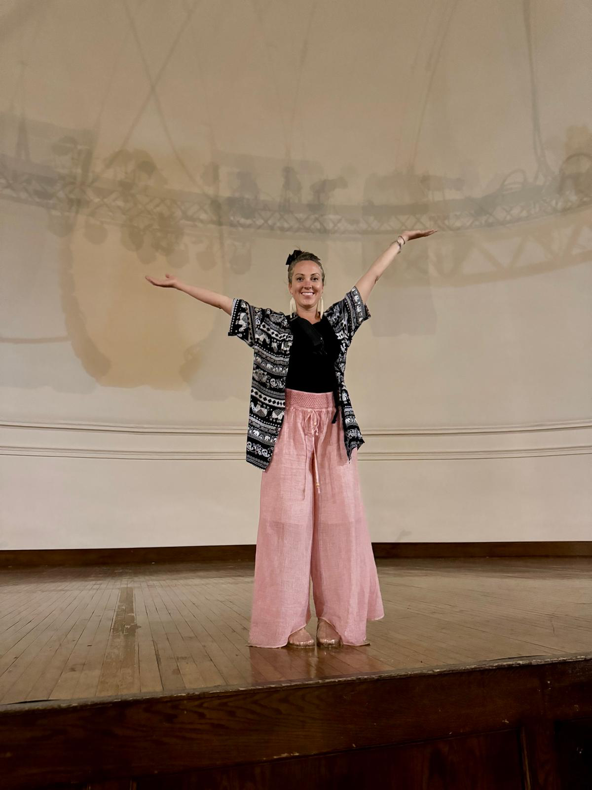 A woman standing on a stage with her arms outstretched, smiling, in front of a large, curved, ceiling. The ceiling has a shadow of stage lights and equipment cast on it.