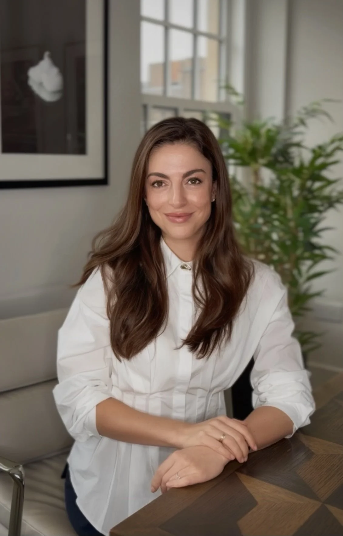A woman with long brown hair, wearing a white shirt, sitting at a wooden table in a bright room with large windows, a colorful framed artwork on the wall, and a green plant in the background.